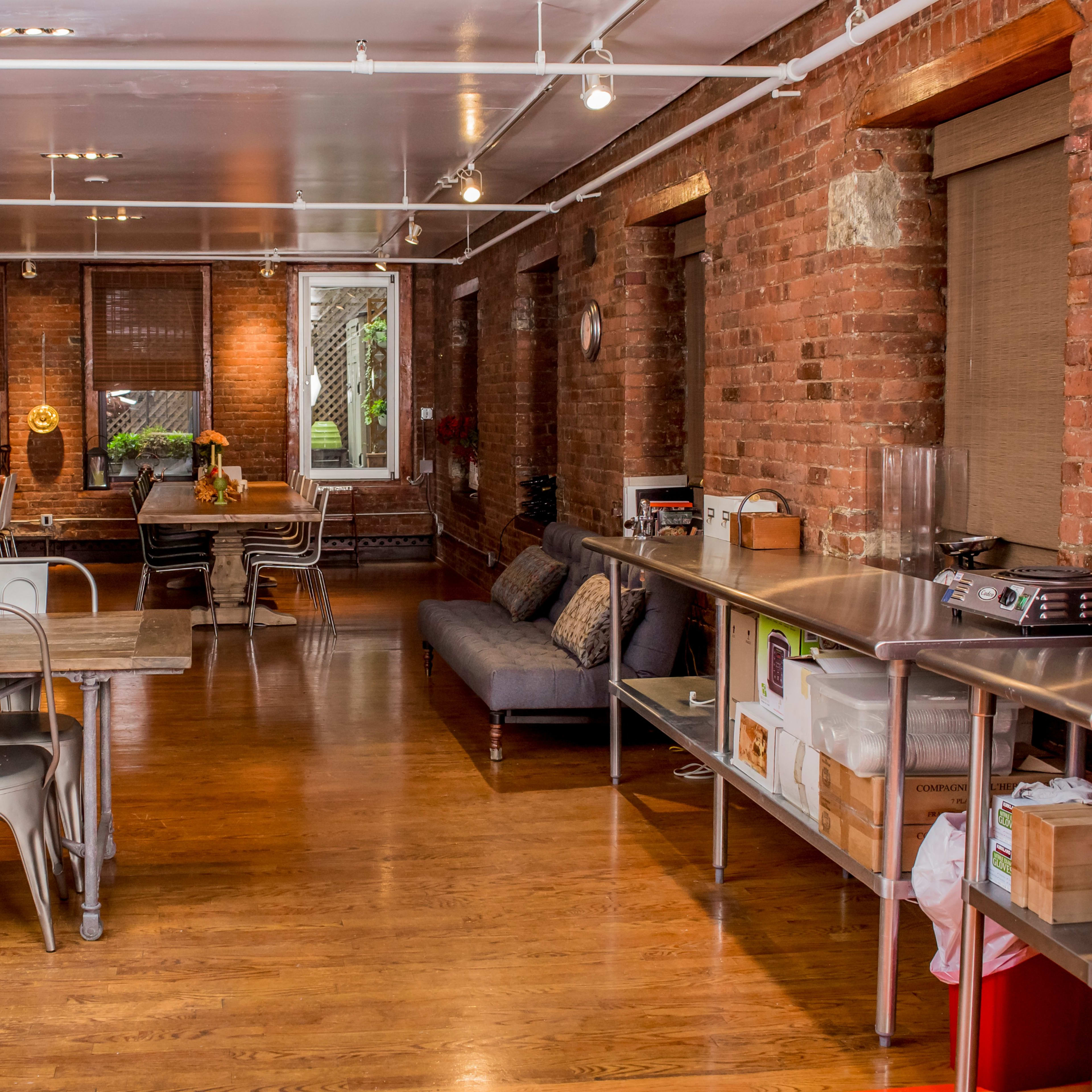 A spacious kitchen area with brick walls, wooden floors, stainless steel counters, and seating arranged around a central space.