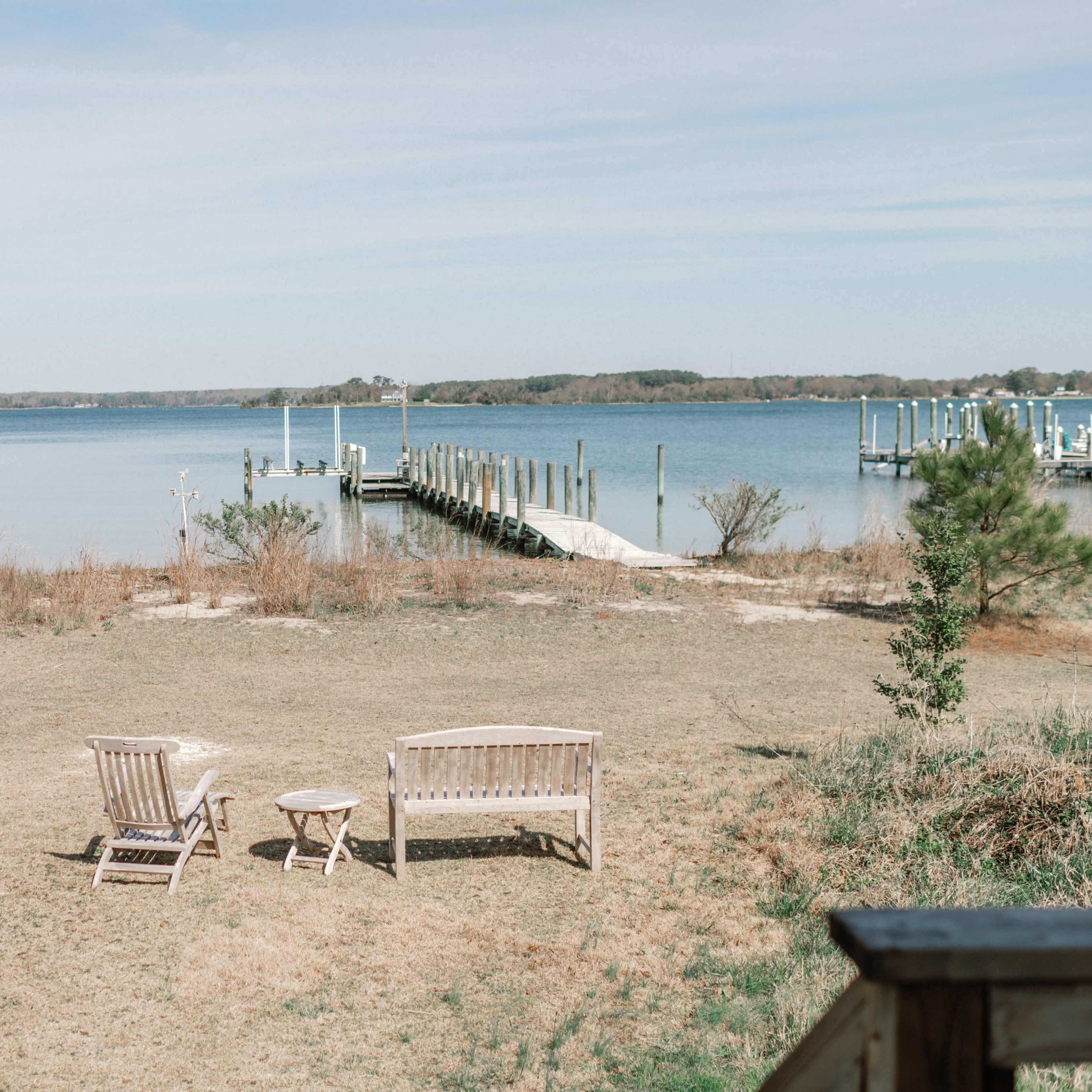 Two wooden chairs and a small round table on a grass area overlooking a calm body of water and a dock.