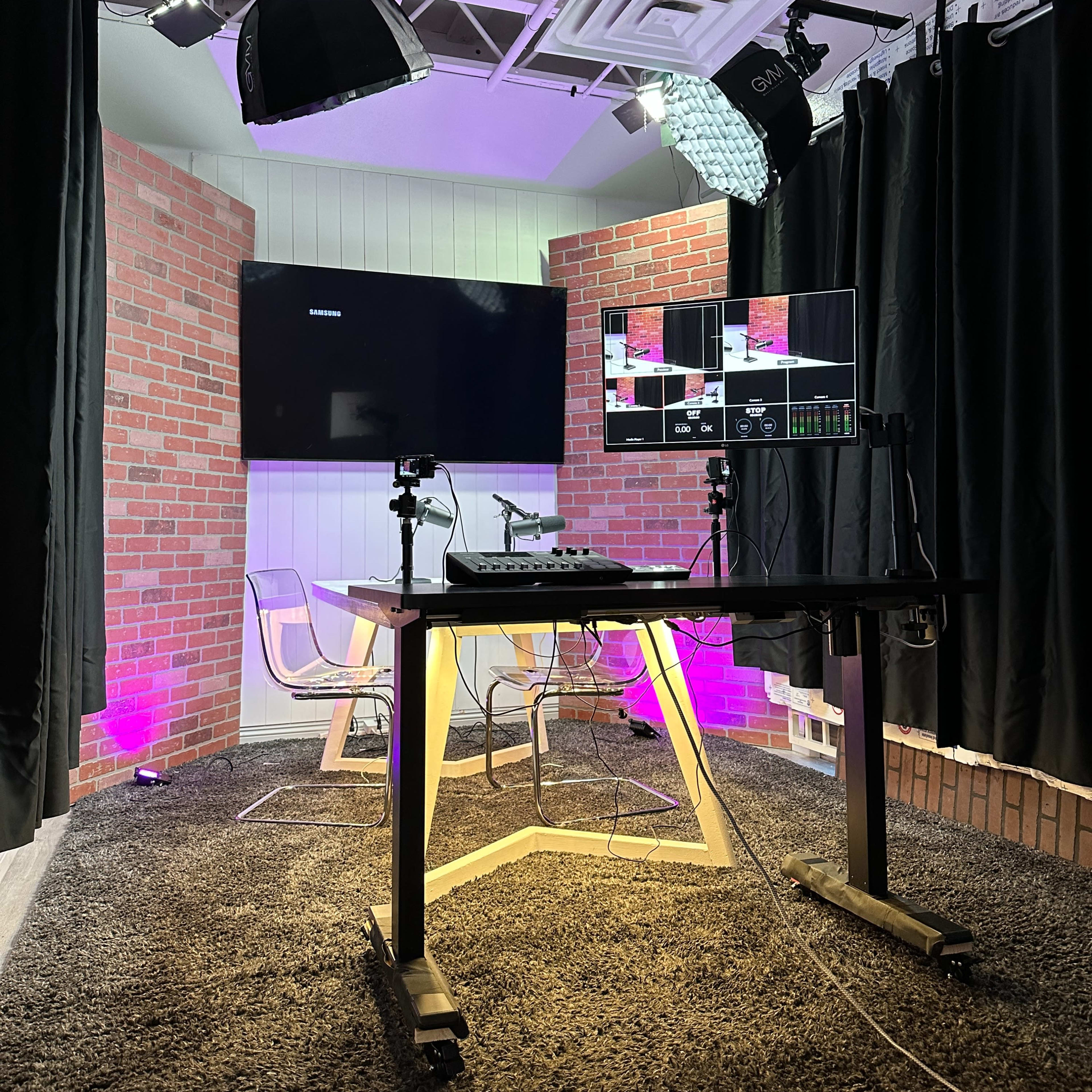 A video production studio with a desk, multiple screens, and brick wall accents, illuminated by soft lighting.