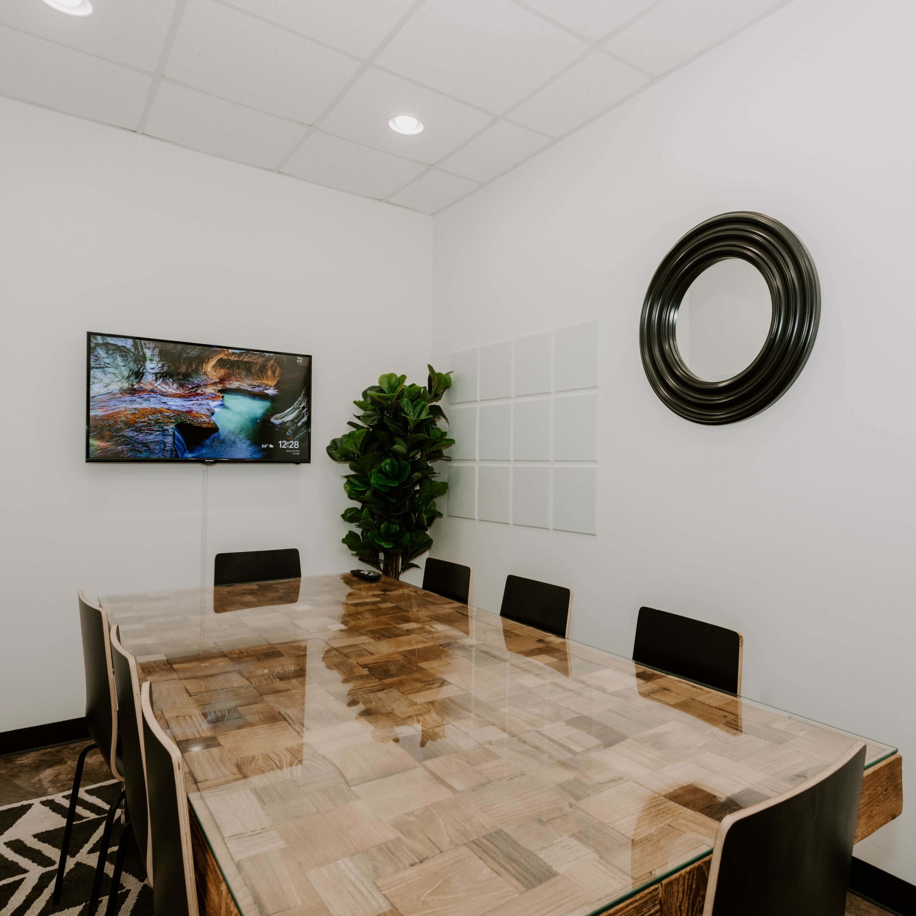 A meeting room features a wooden table surrounded by black chairs, a wall-mounted TV, and a decorative plant.