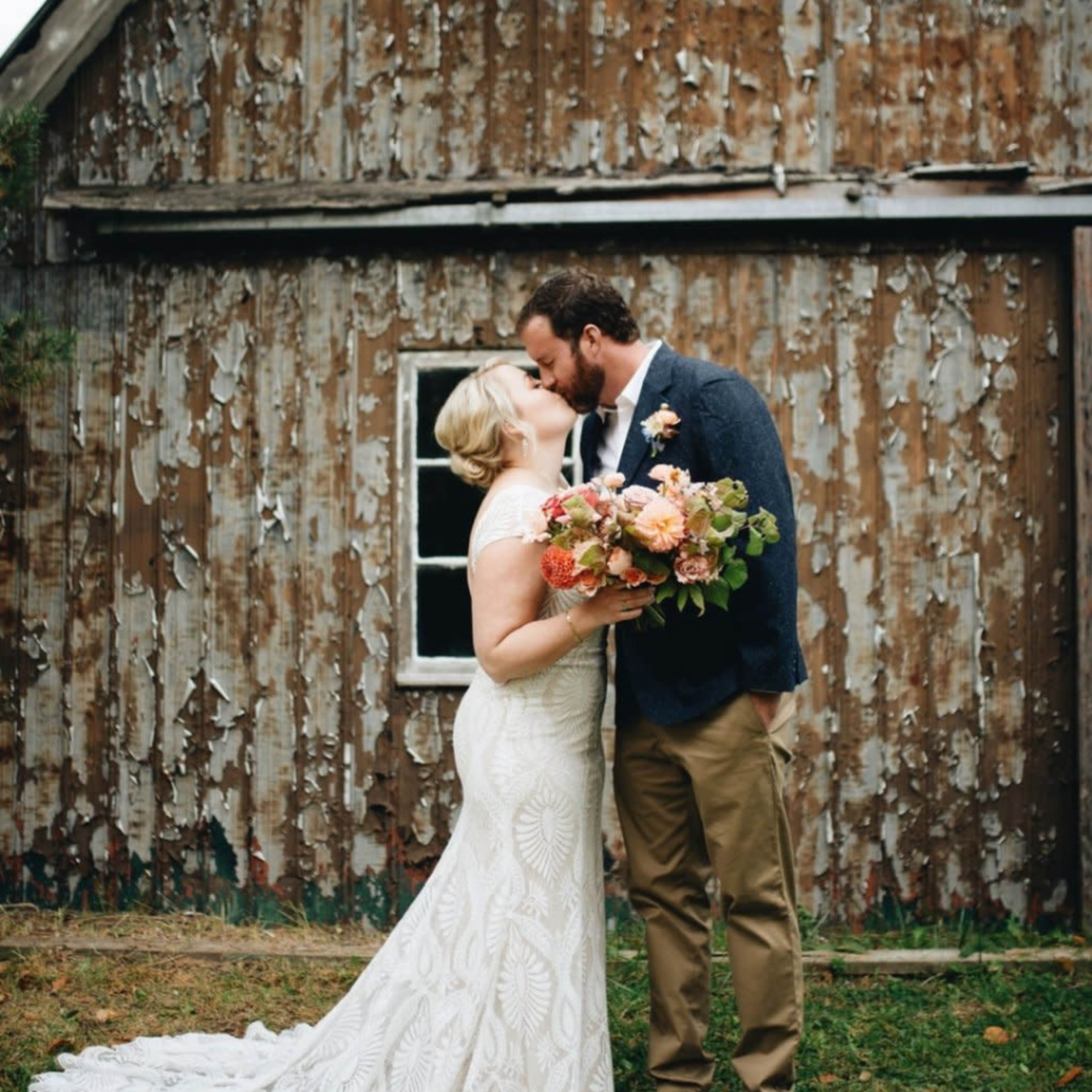 A bride in a lace wedding dress kisses a groom in a suit outside a rustic, weathered building.