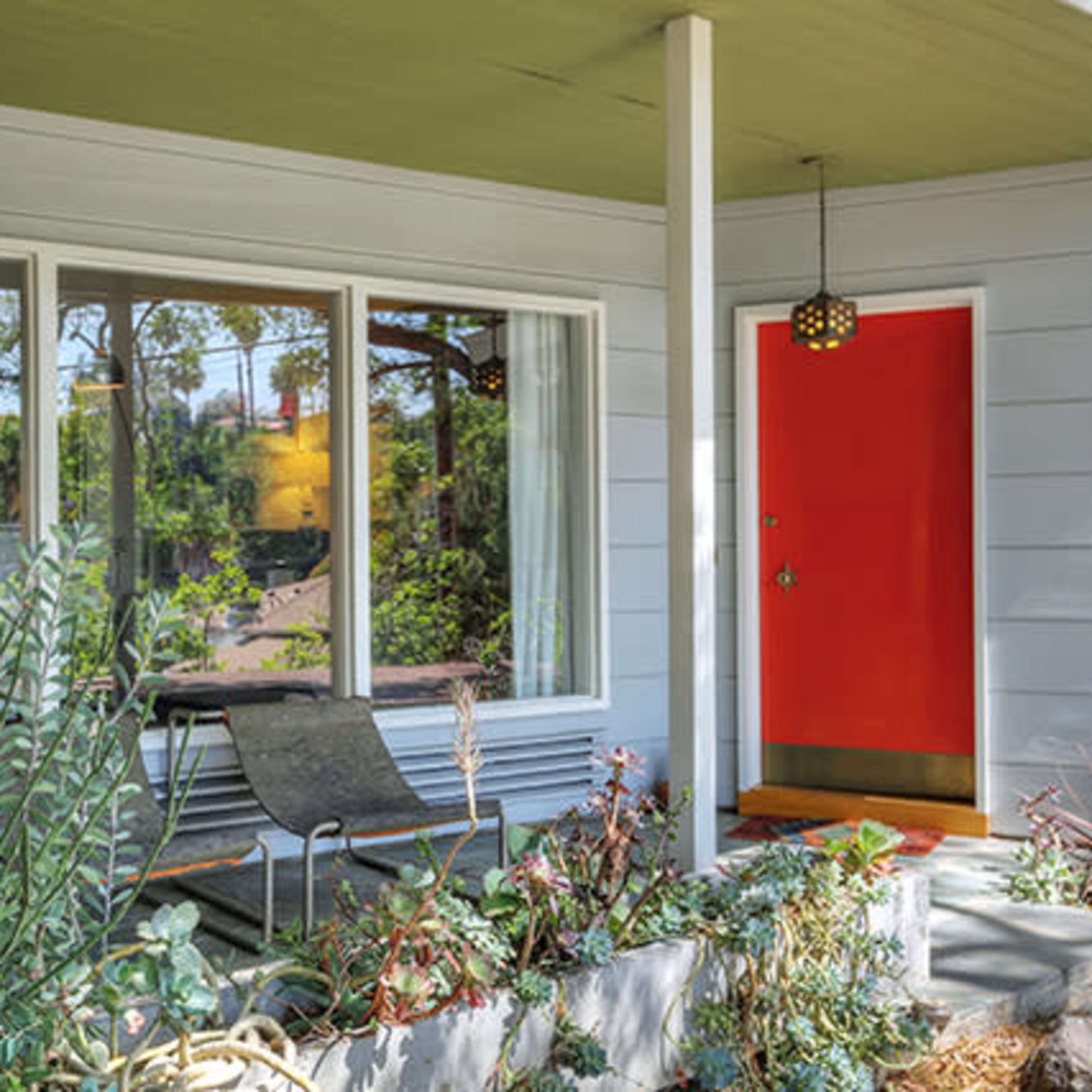 A modern house facade with a bright red door, large front windows, and a small seating area surrounded by plants.
