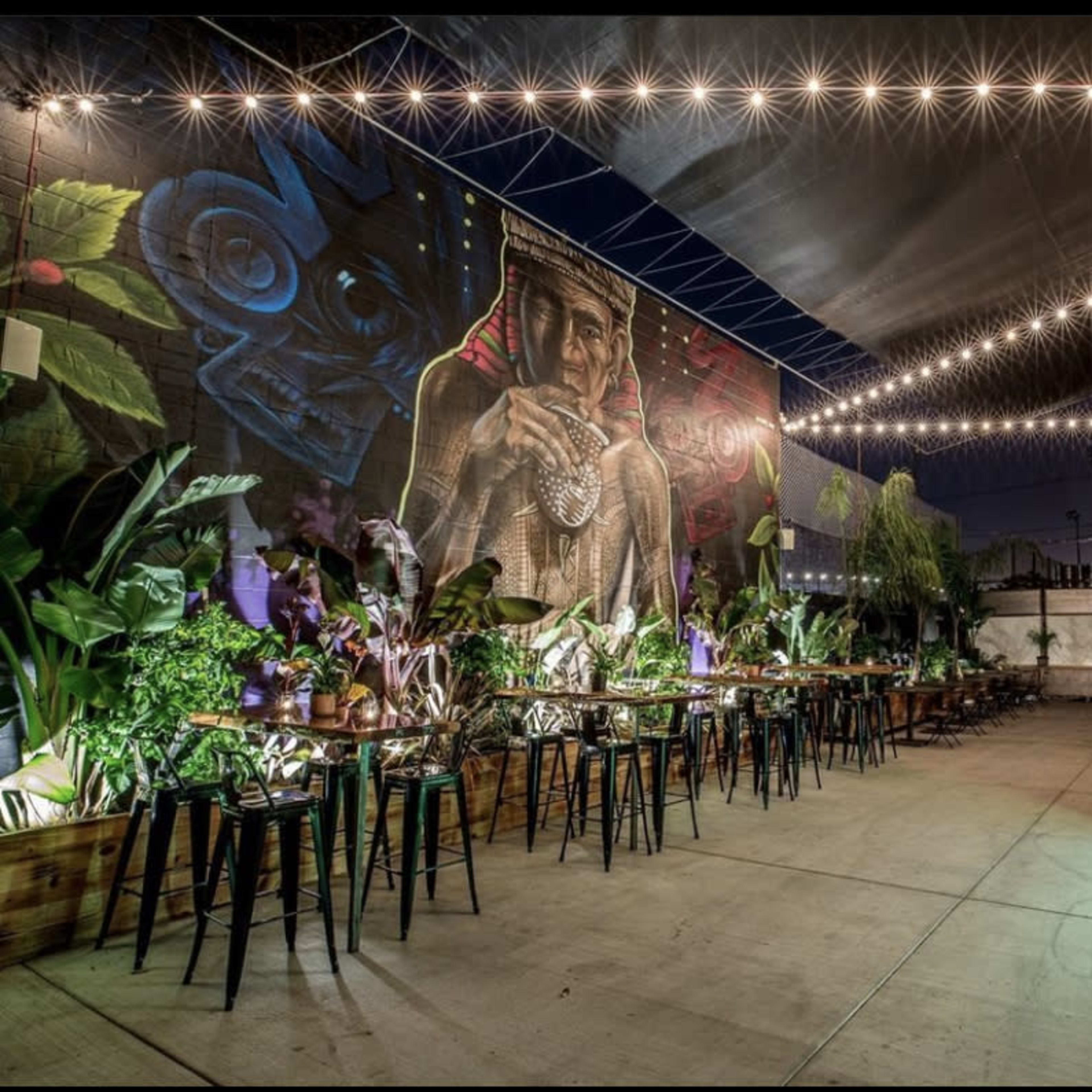 A spacious outdoor dining area featuring string lights overhead, tables with black stools, and a vibrant mural on the back wall.