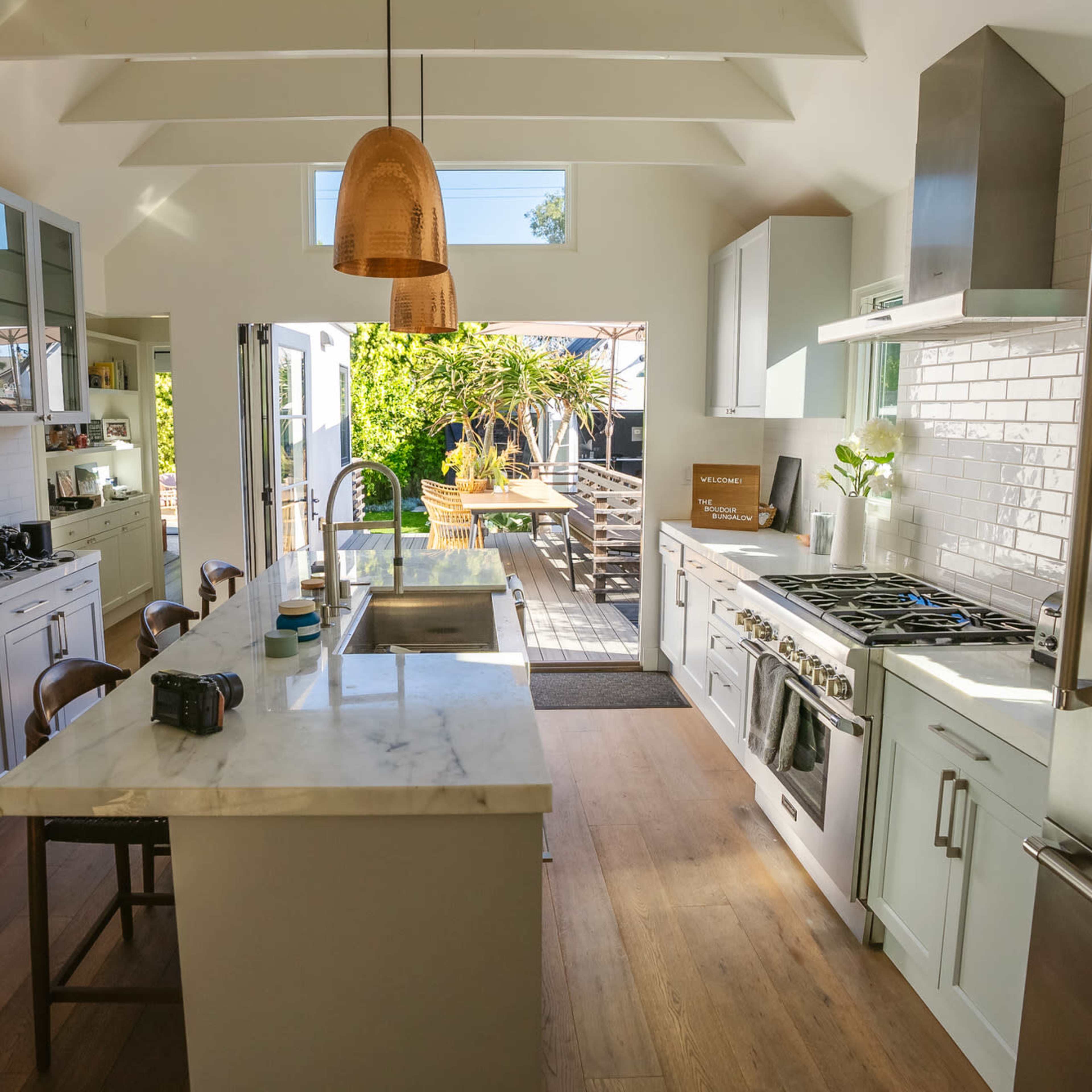 A spacious kitchen features a central island with a marble countertop, stainless steel appliances, and large windows that open to an outdoor dining area.