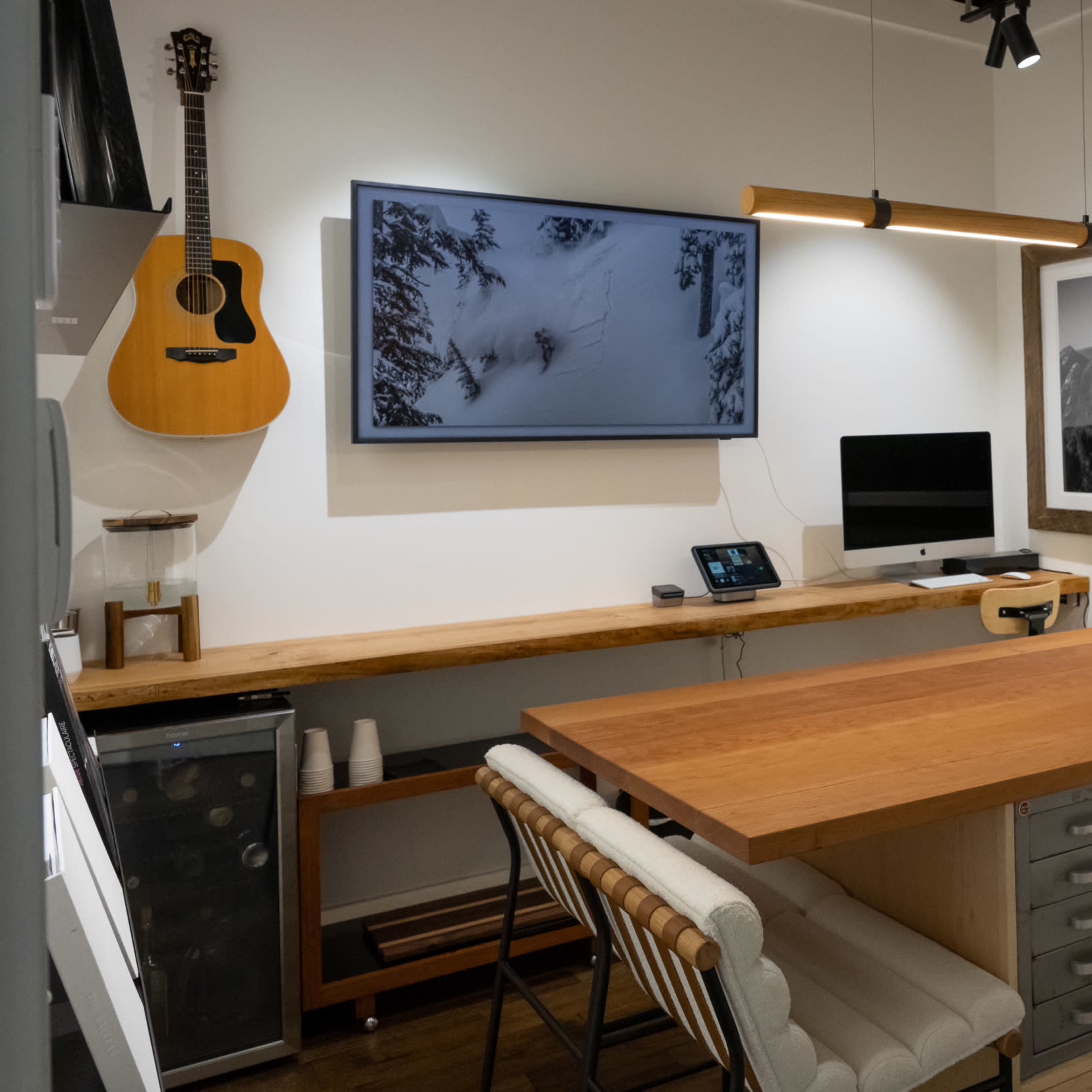 A modern workspace with a wooden table, two monitors, an acoustic guitar hanging on the wall, and a large photograph of mountains.