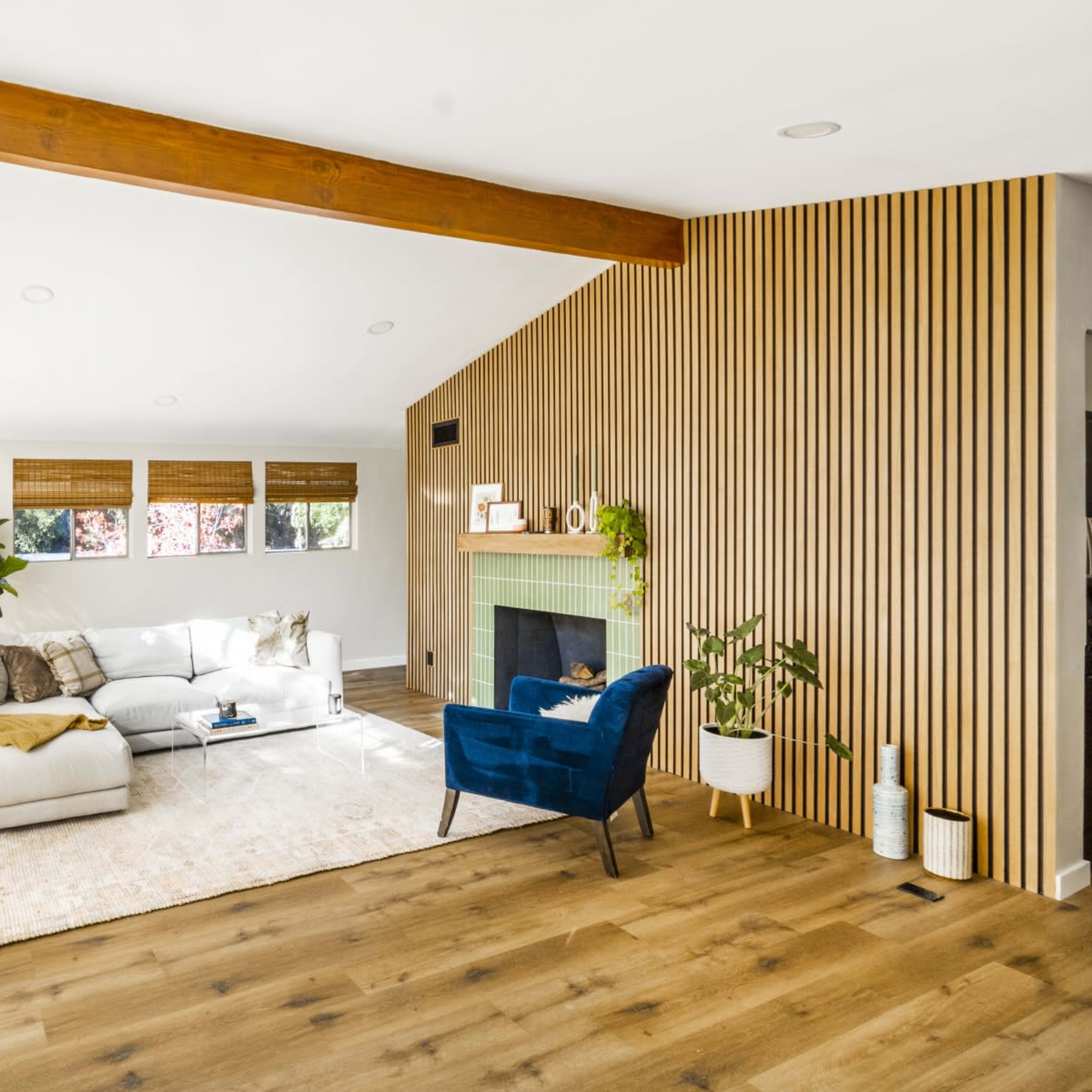 A modern living room features a light-colored sectional sofa, a blue armchair, a plant, and a fireplace, with large windows allowing natural light to enter.