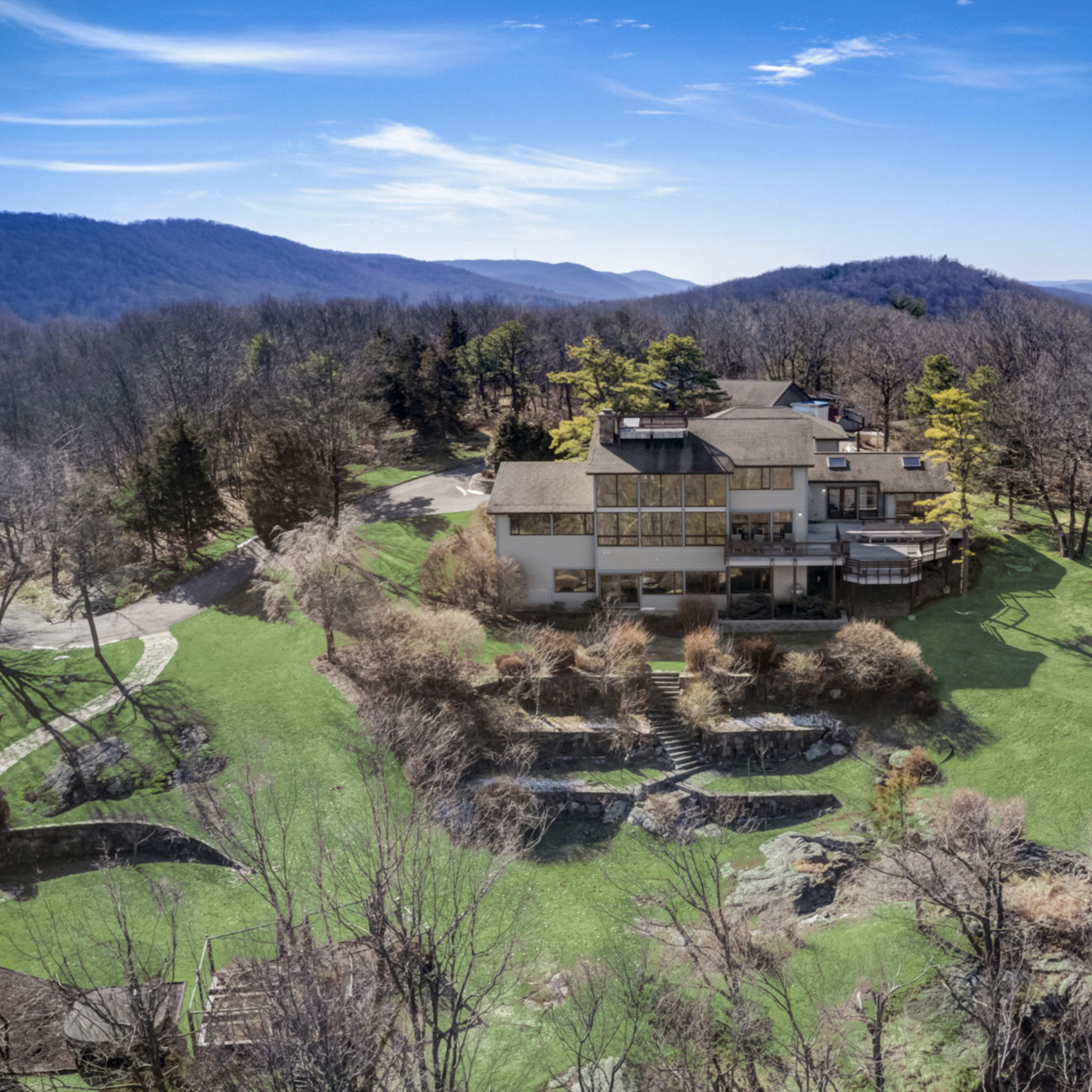 A large, modern house sits on a green hillside surrounded by sparse trees and distant mountains under a clear blue sky.