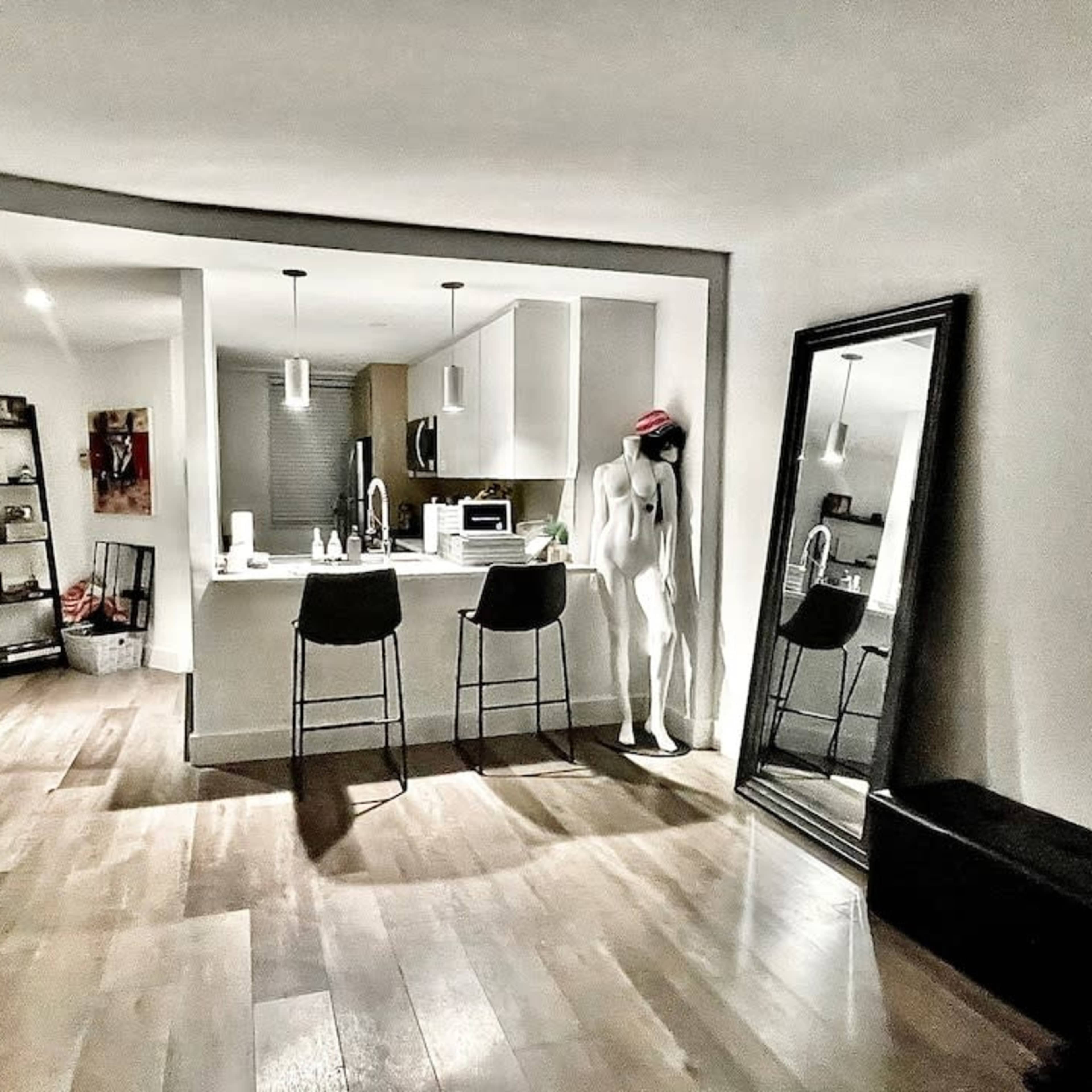 A modern kitchen with a central island, bar stools, a mannequin wearing a red hat, and a large mirror reflecting the surroundings.