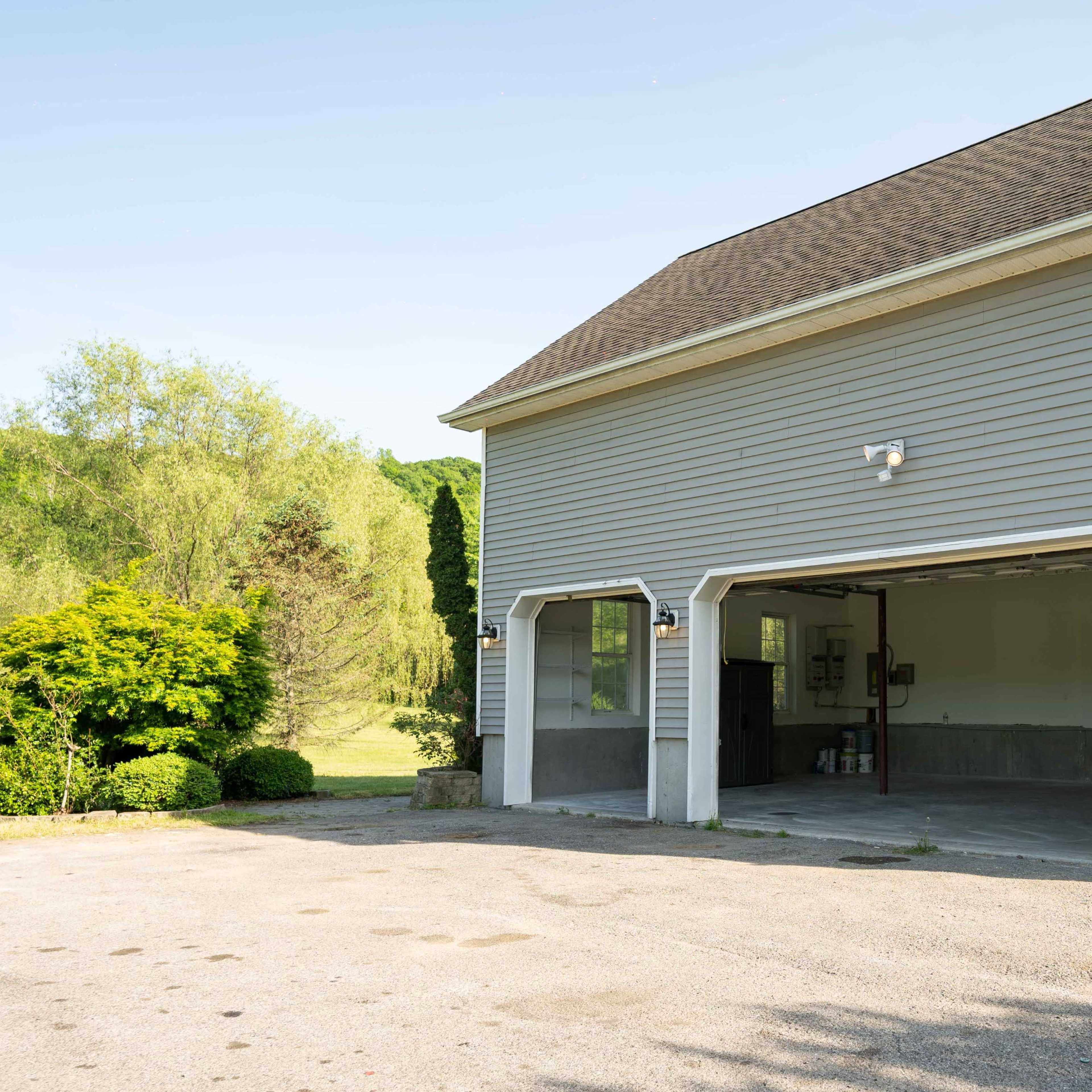 A gray, two-story building with a garage featuring two open doors and a white entrance door, situated next to a well-maintained gravel area and lush greenery.