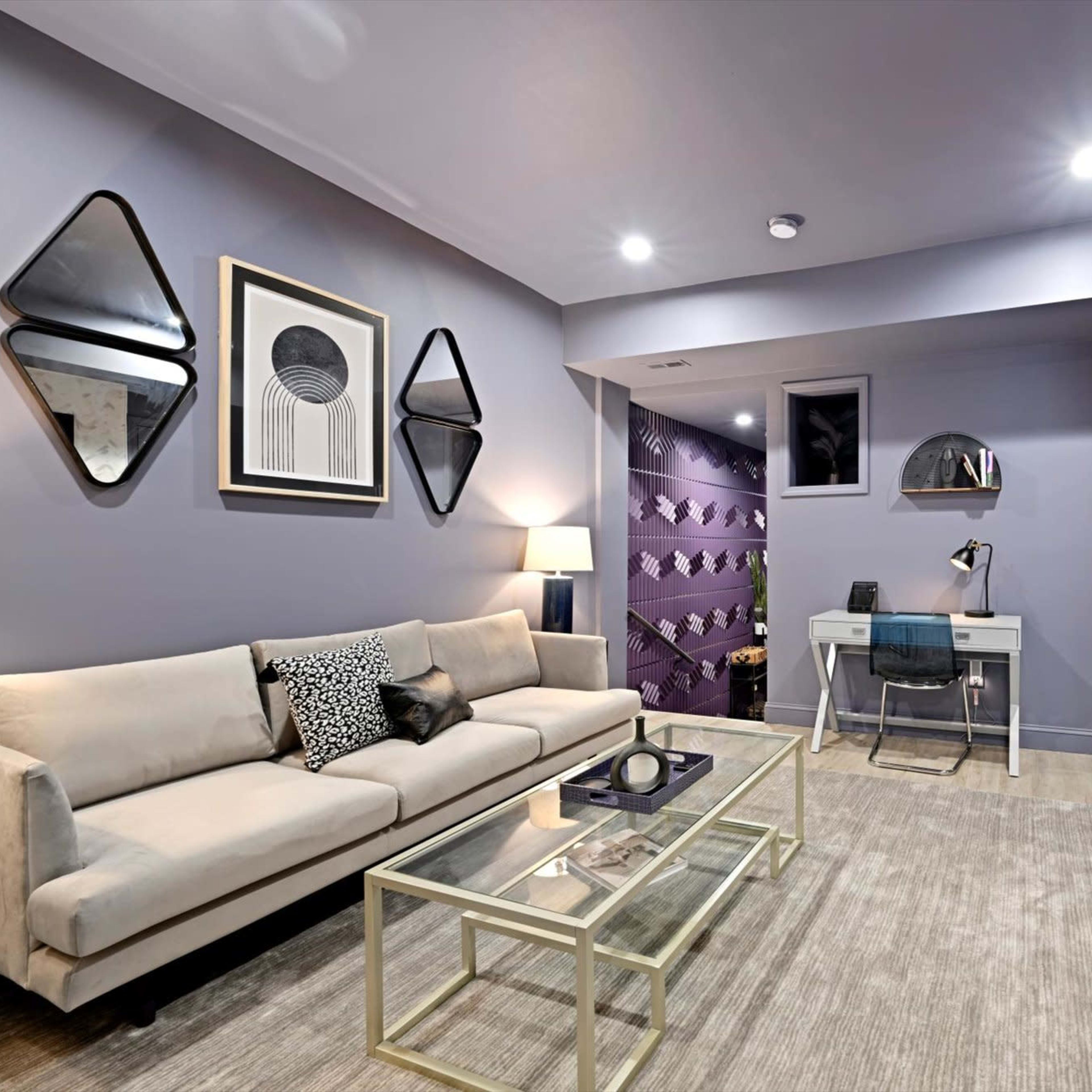 A modern living room features a light-colored sofa, glass coffee table, and a sleek fireplace, with decorative mirrors and a desk set against stylish wall designs.