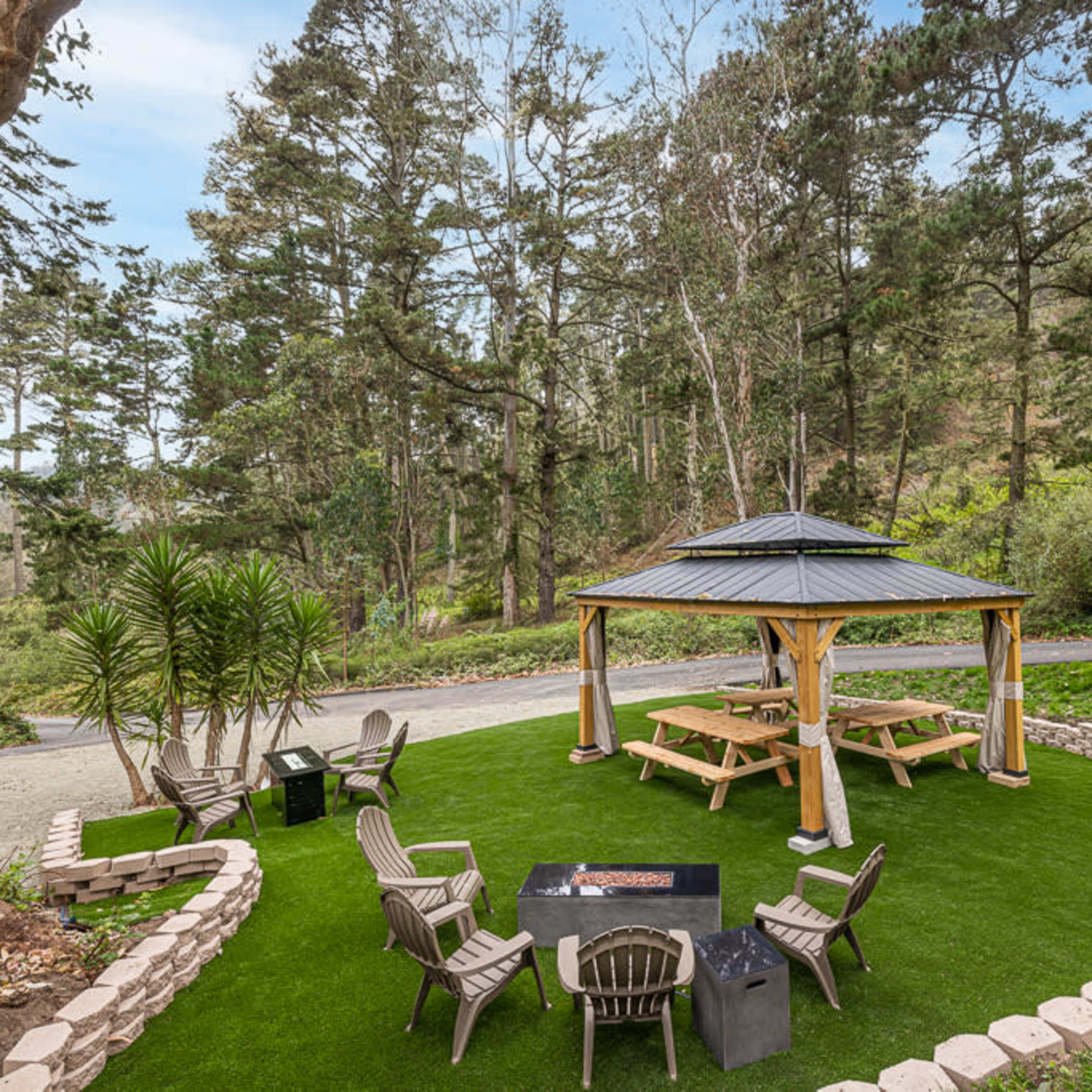 A gazebo with picnic tables and seating surrounded by landscaped grass and trees in a wooded area.