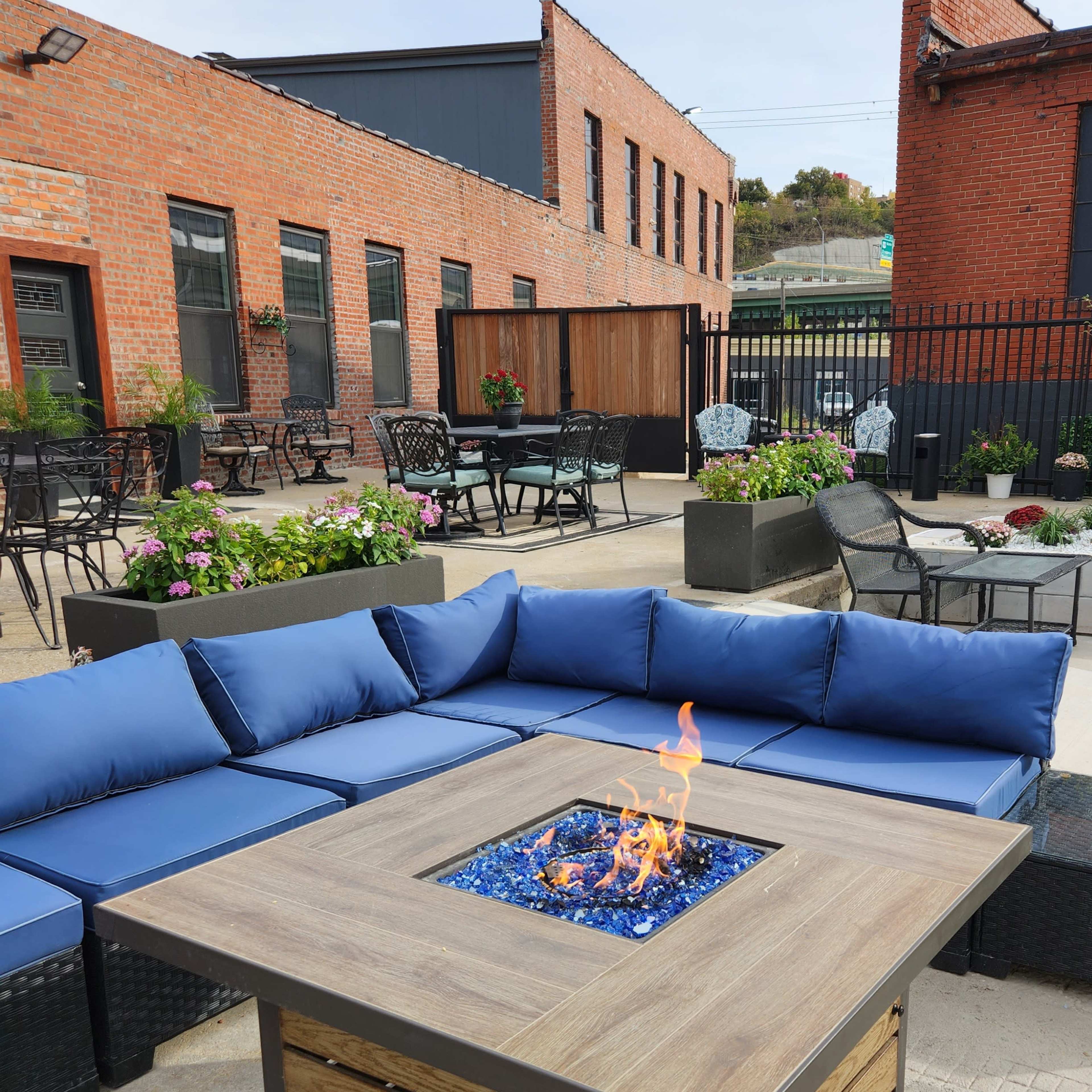 A square fire pit is centered within a wooden table on a patio surrounded by blue couches and potted plants.