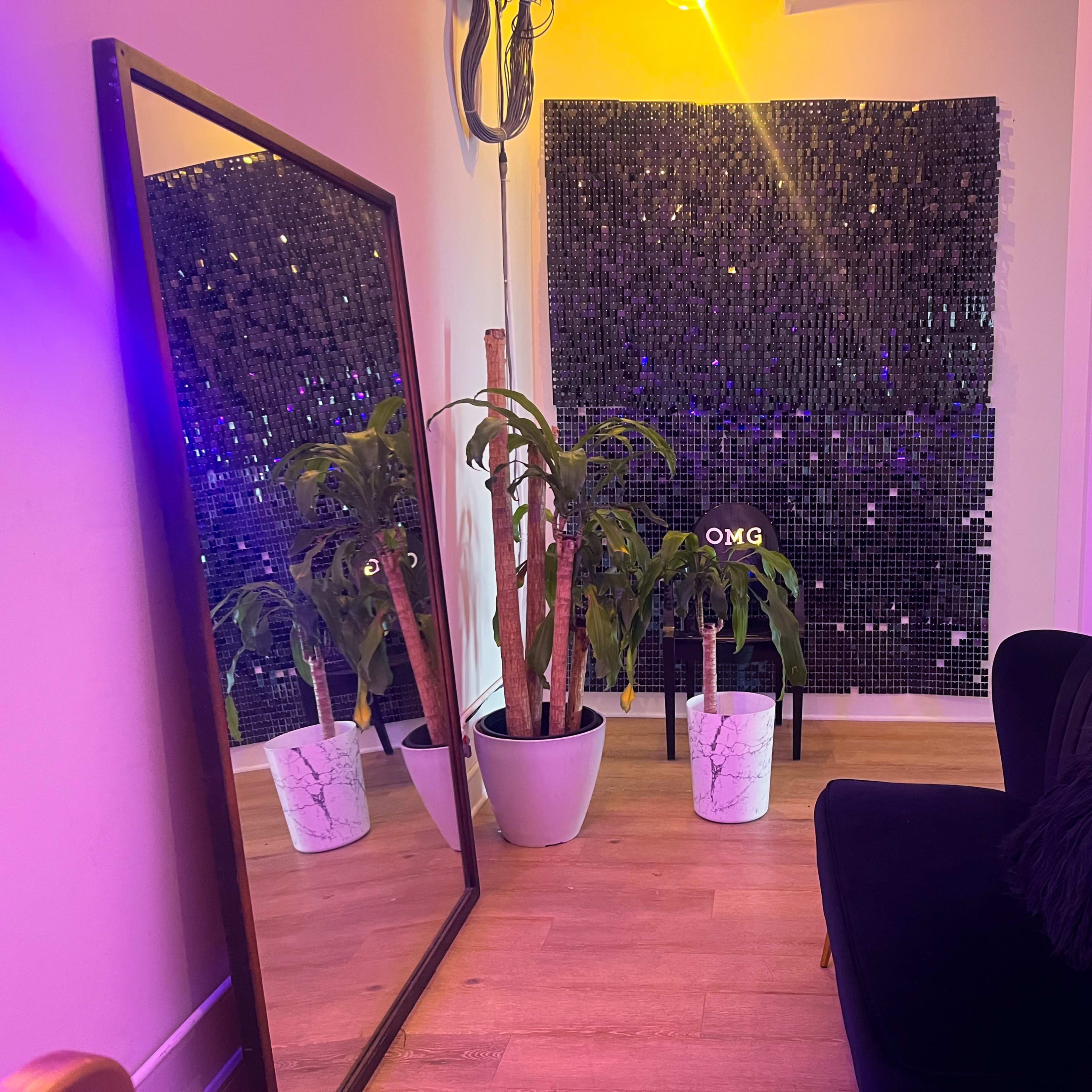 A small room features a large mirror, a black sofa, a potted plant, and a decorative wall made of reflective tiles.