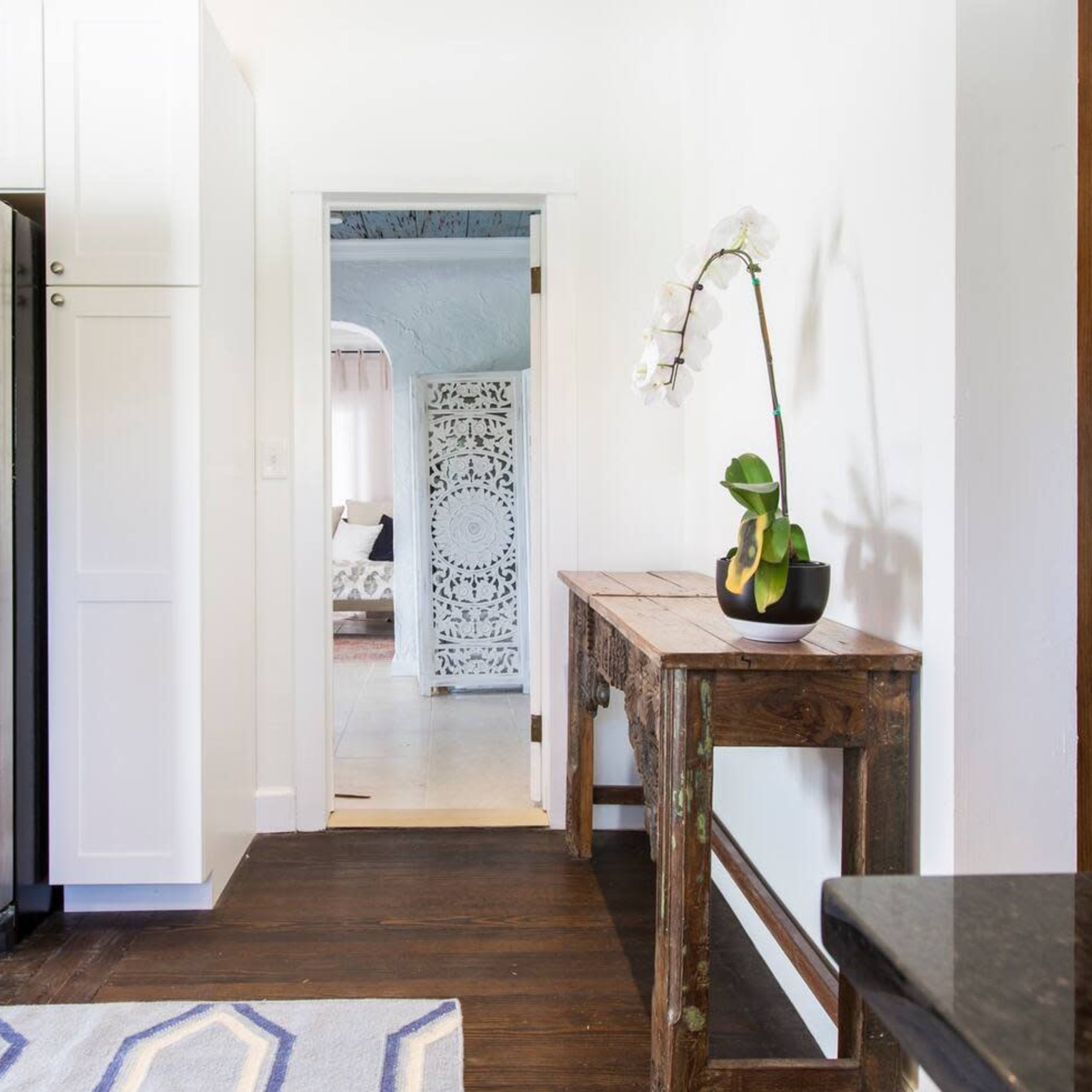 A kitchen area with a stainless steel refrigerator, a wooden console table with a potted orchid, and a doorway leading to a living room.