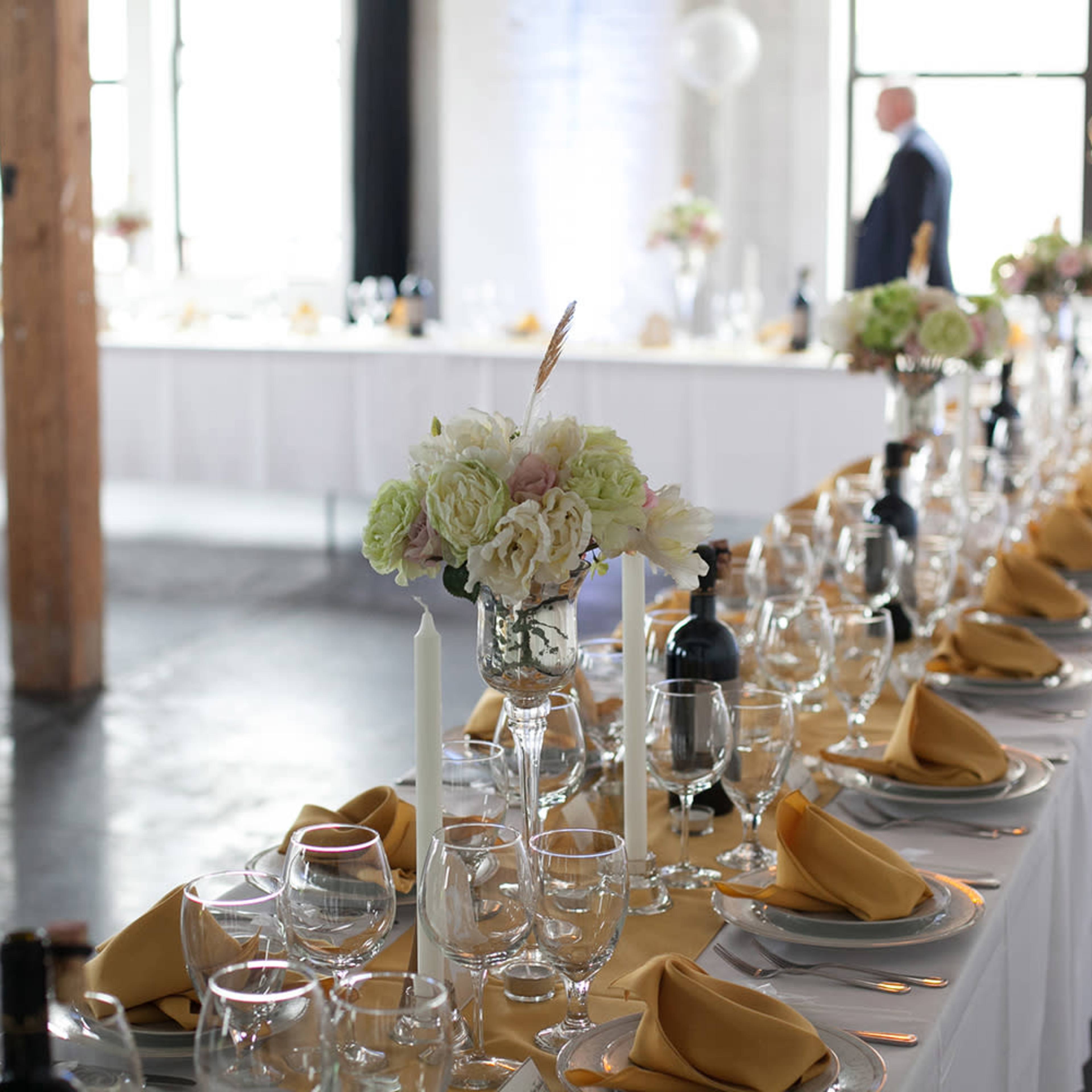 A long dining table is elegantly set with white tablecloths, gold napkins, glassware, and floral centerpieces, in a spacious room with large windows.