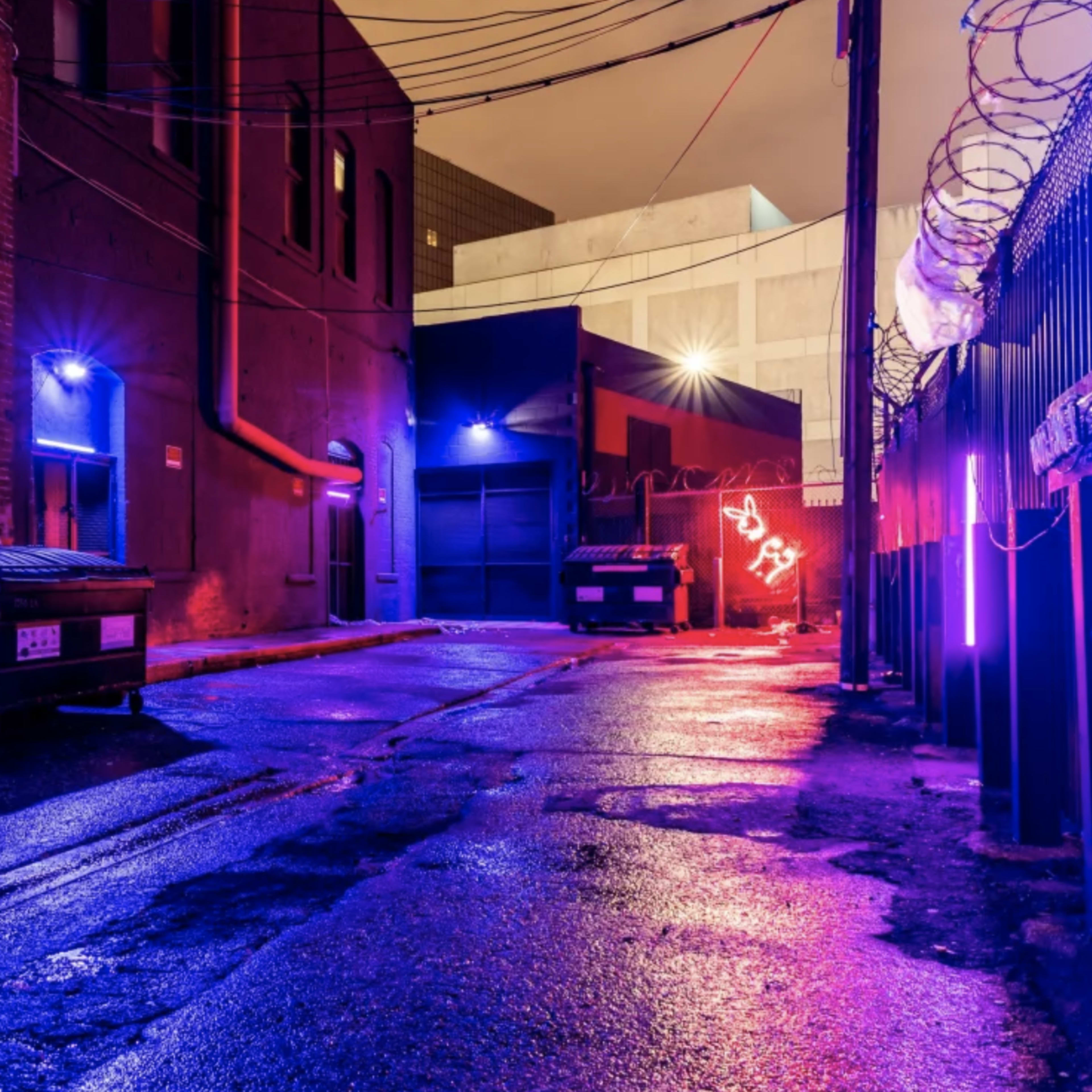 A wet alleyway at night, featuring a dumpster, a parked car, and illuminated signs casting colorful lights along the brick walls.