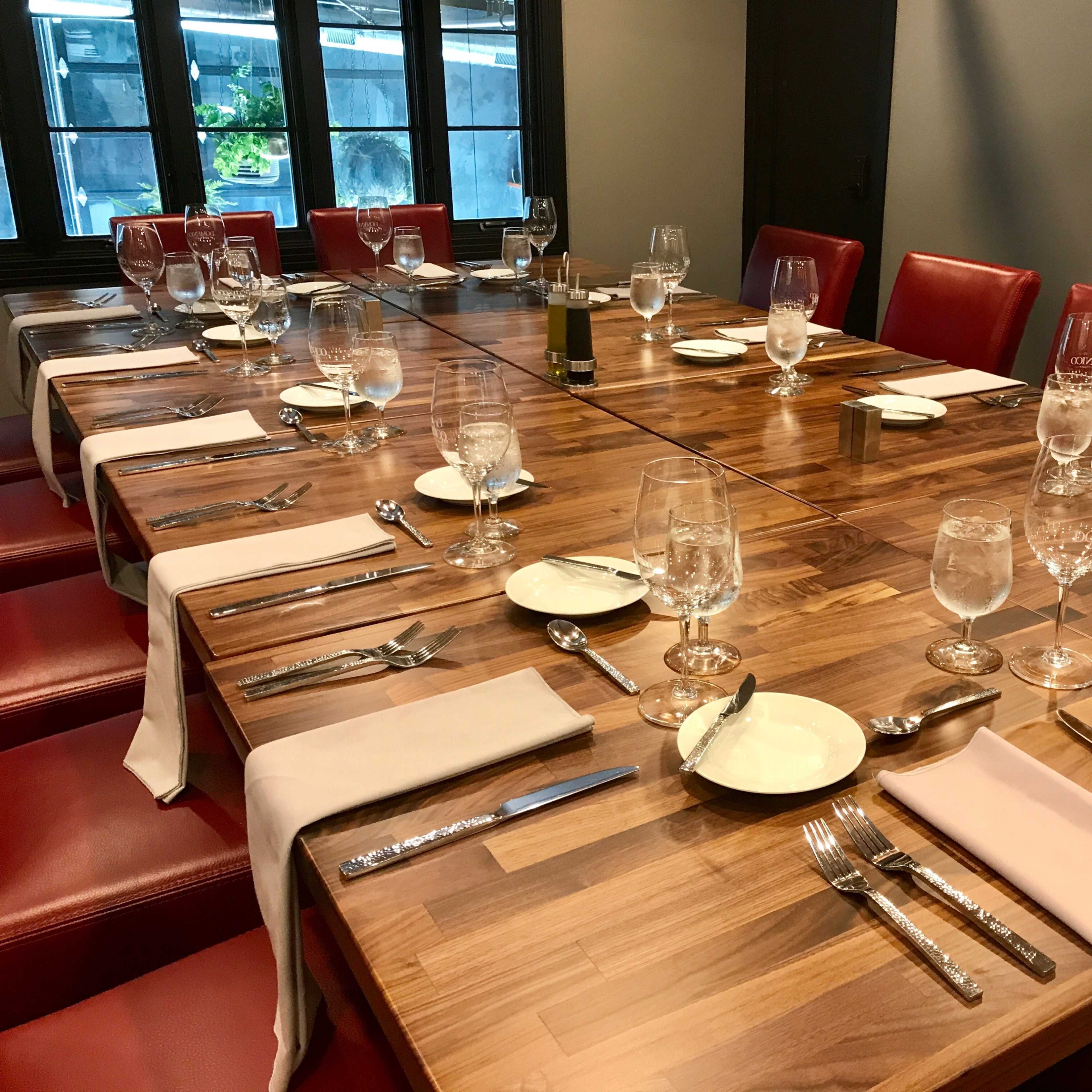 A long wooden dining table is set with plates, glasses, and utensils in a room with red chairs and large windows.