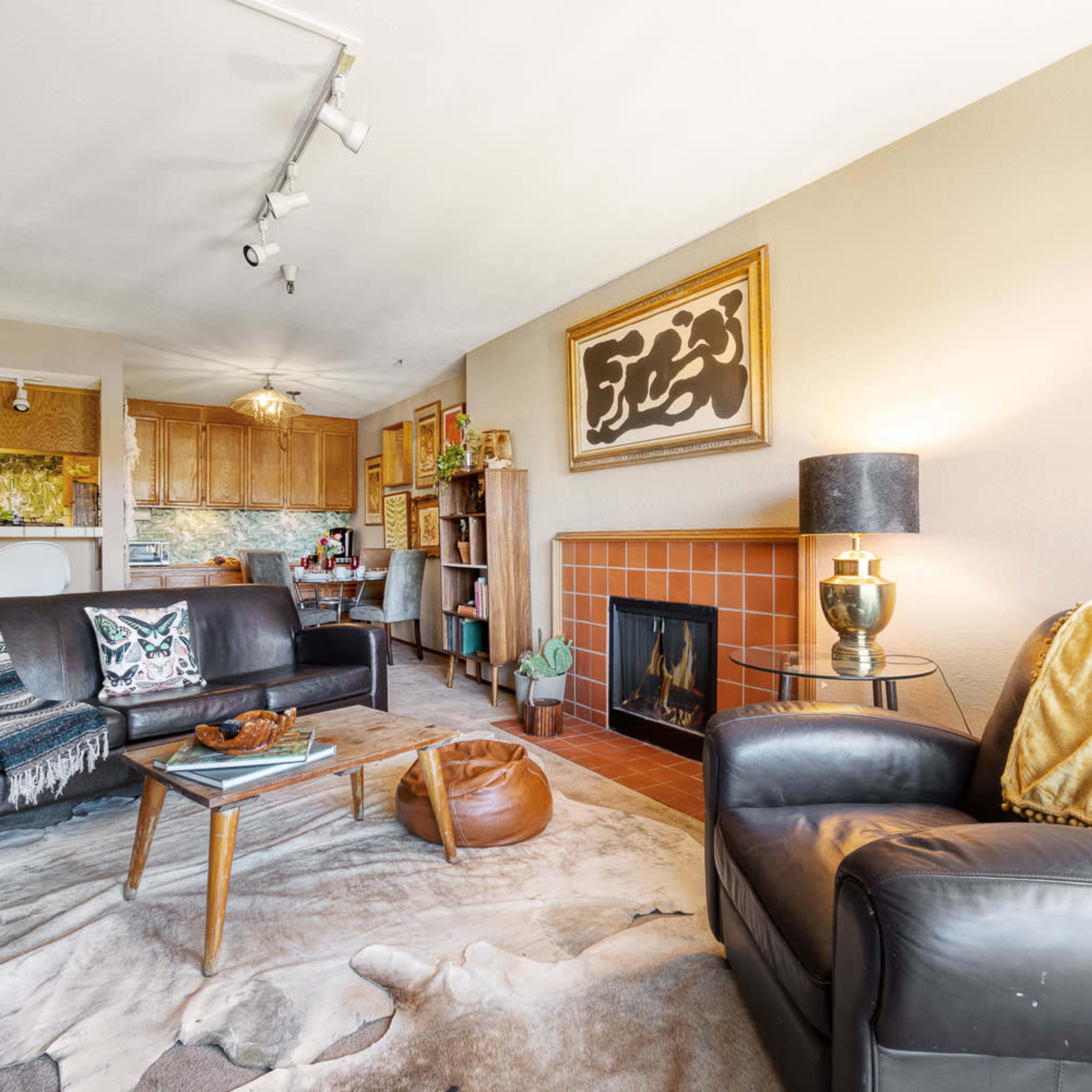 A cozy living room featuring a fireplace, a leather armchair, a coffee table, and a kitchen in the background.