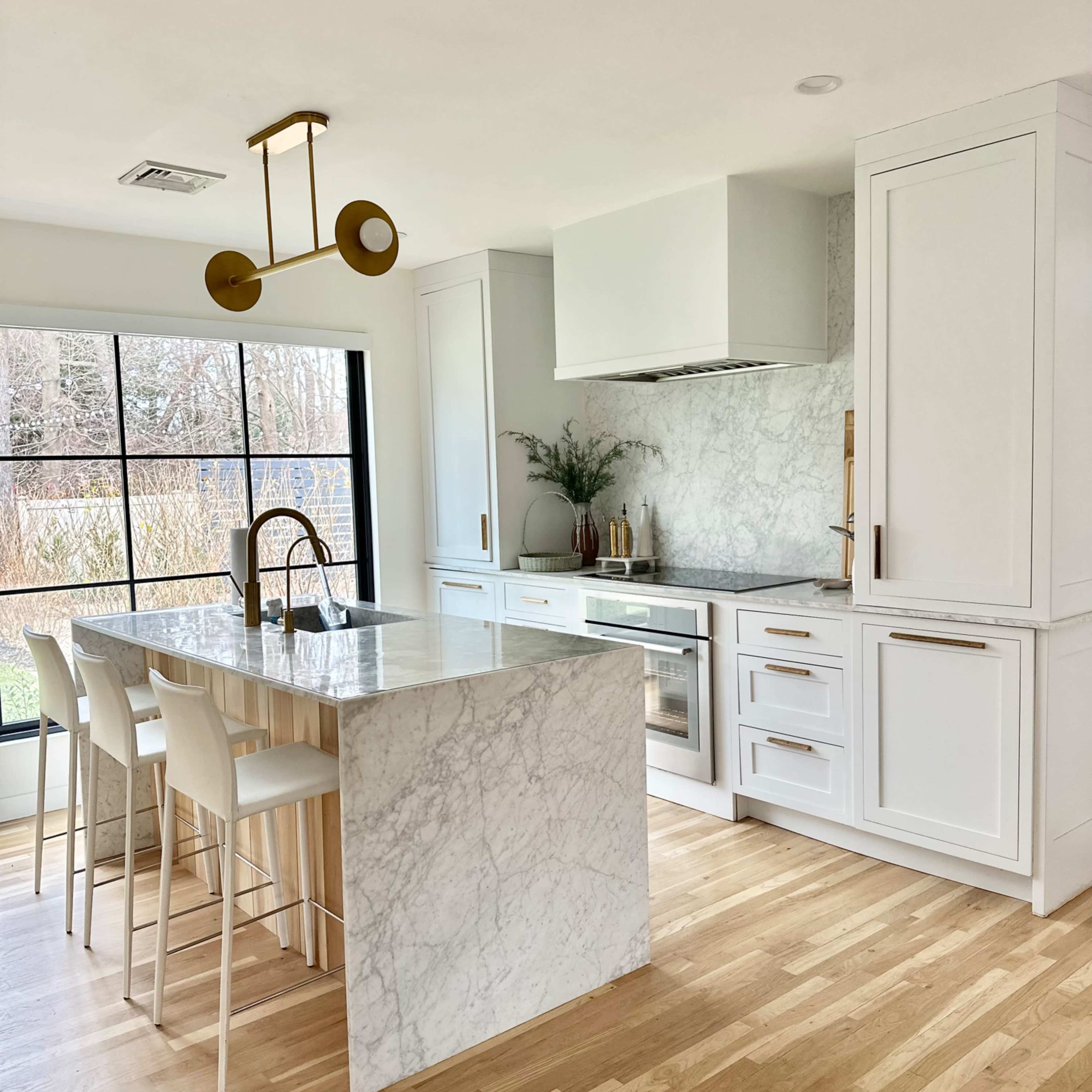 A modern kitchen features white cabinetry, a large marble island with bar stools, and a window providing natural light.