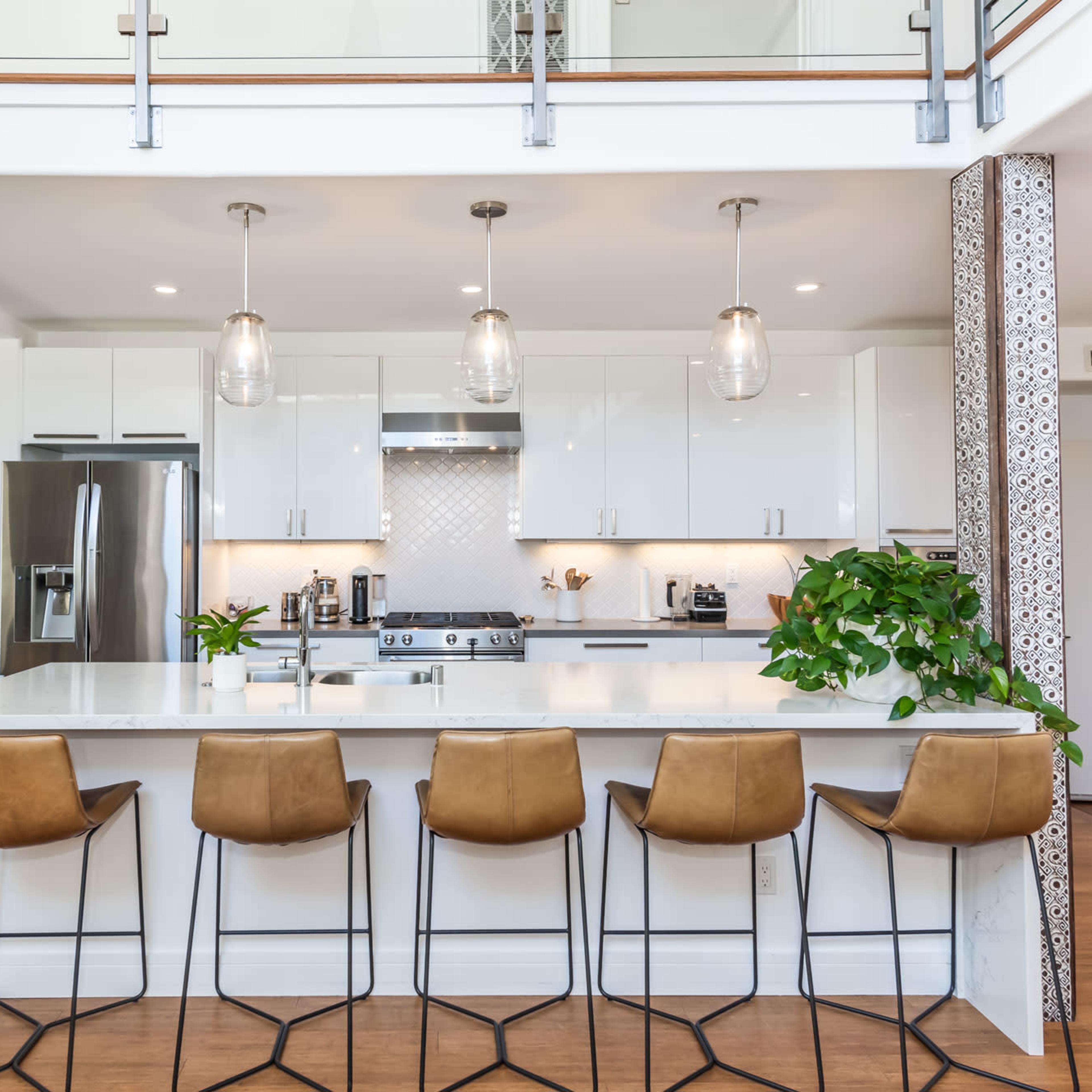 A modern kitchen with a white island counter, three pendant lights, and bar stools, situated in an open floor plan with plants and stainless steel appliances.