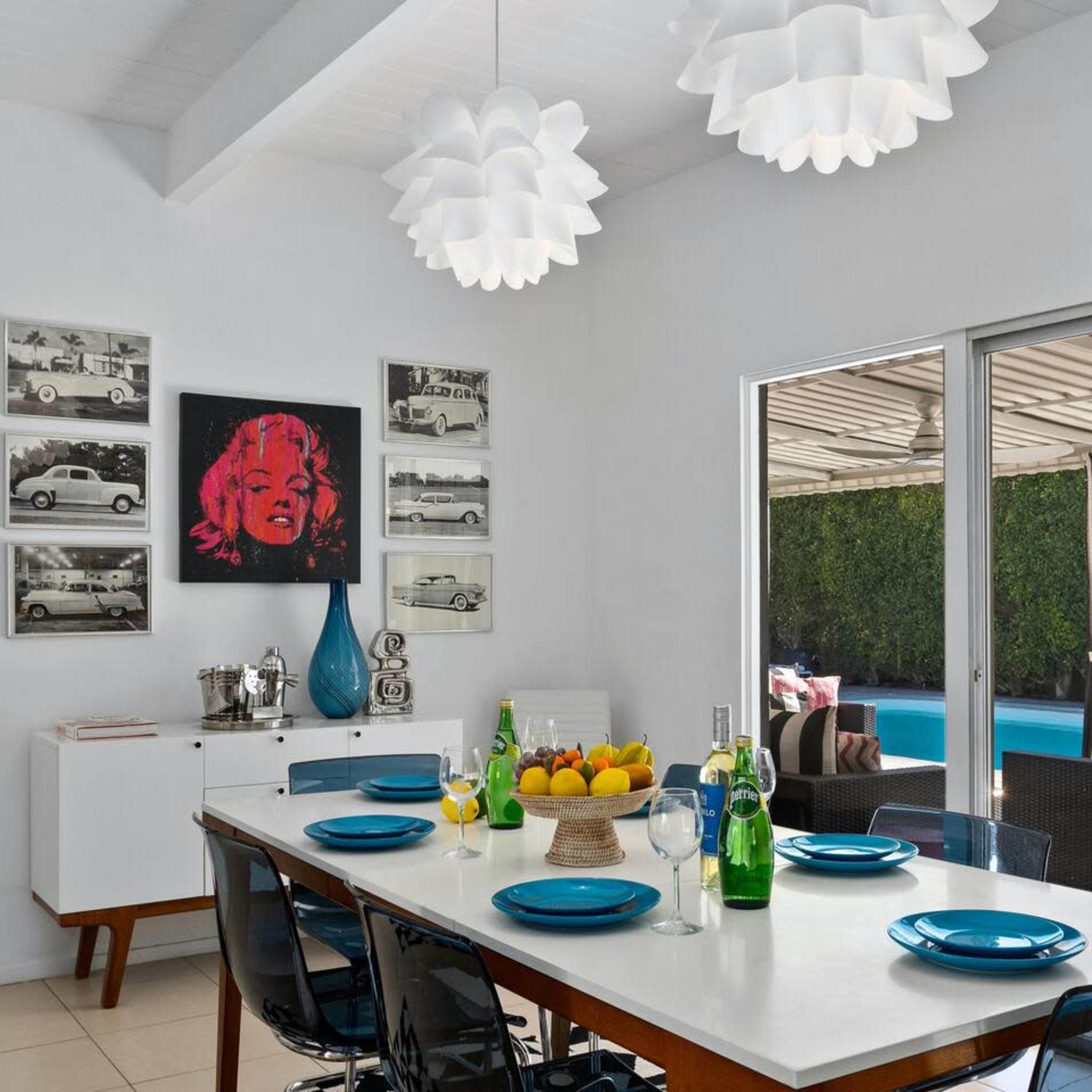 A modern dining area features a white table set with blue plates, surrounded by black chairs, while facing a large window that opens to a poolside patio.