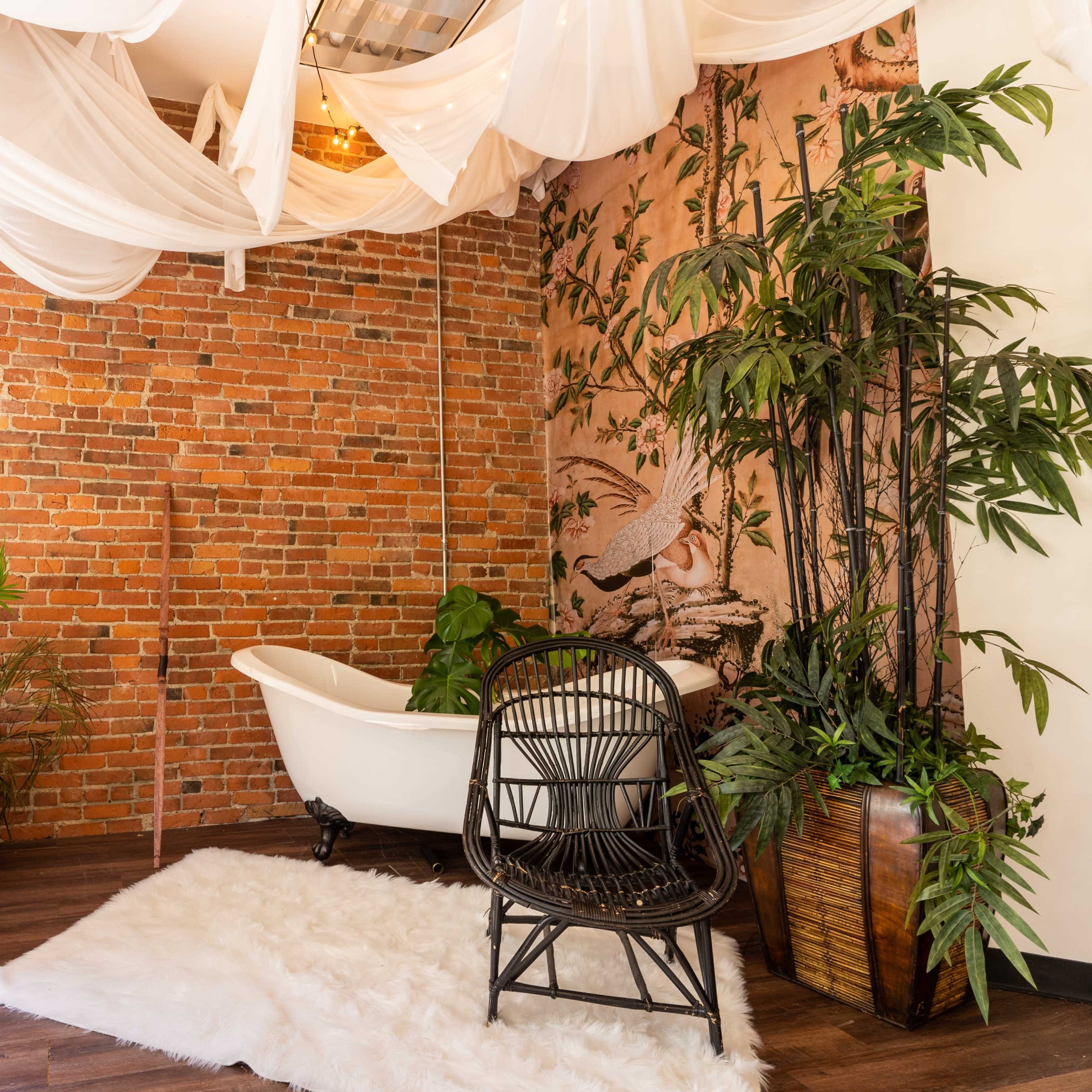 A cozy corner of a room with exposed brick walls, a freestanding bathtub, a wicker chair, and greenery, accented by string lights and draped fabric overhead.