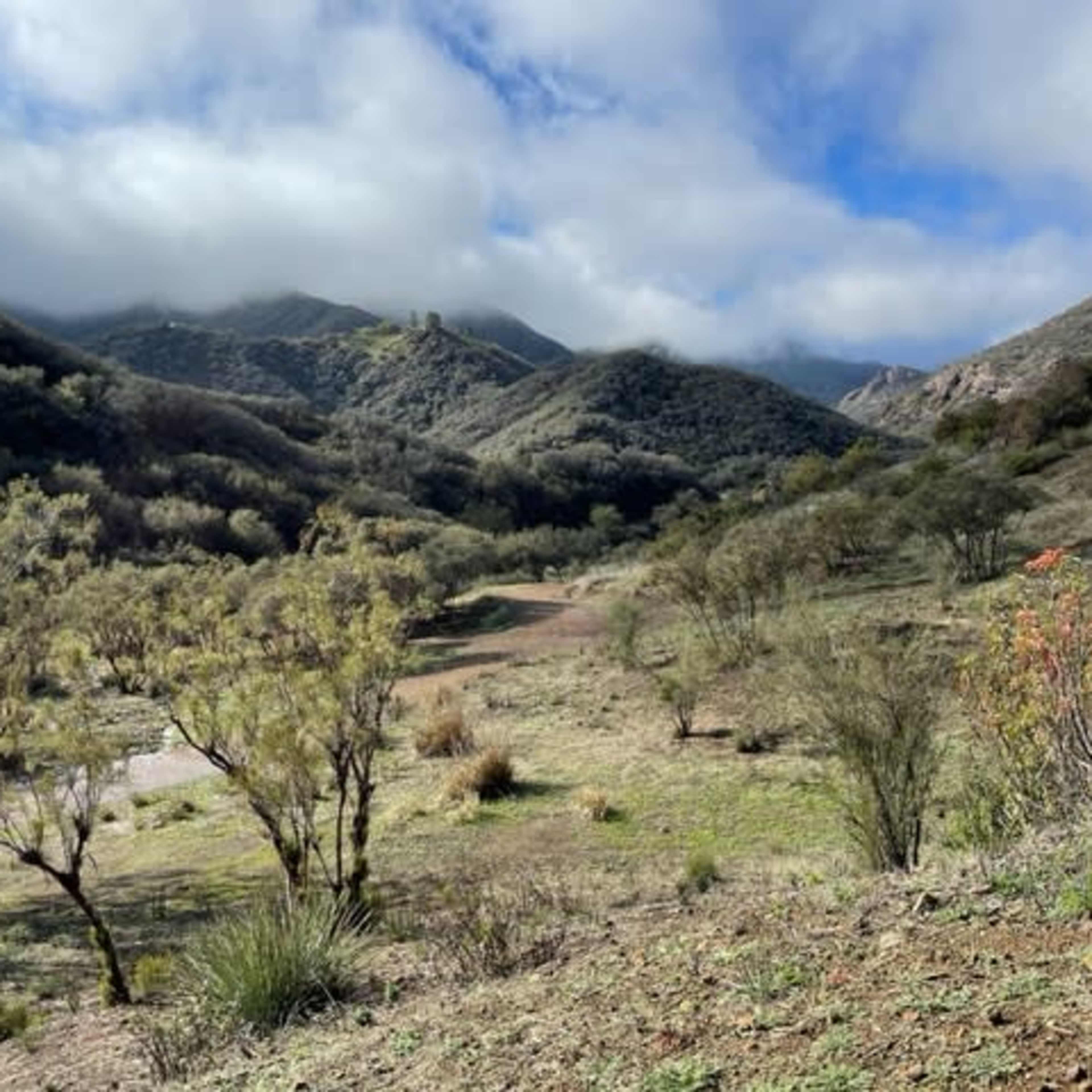 A winding dirt path leads through a hilly landscape covered with sparse vegetation under a cloudy sky.