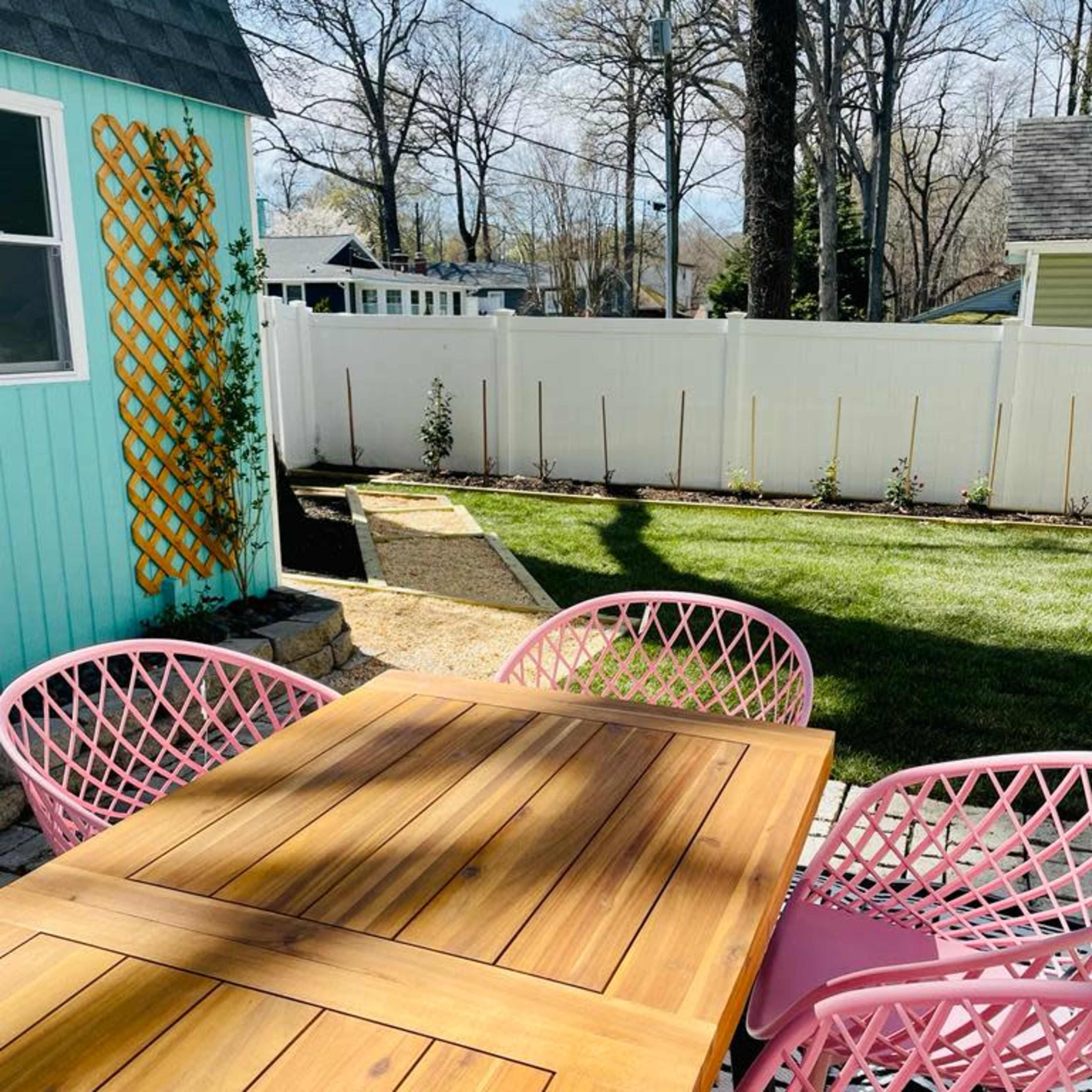 A wooden table with pink chairs is set in a backyard featuring a green lawn and a white fence.