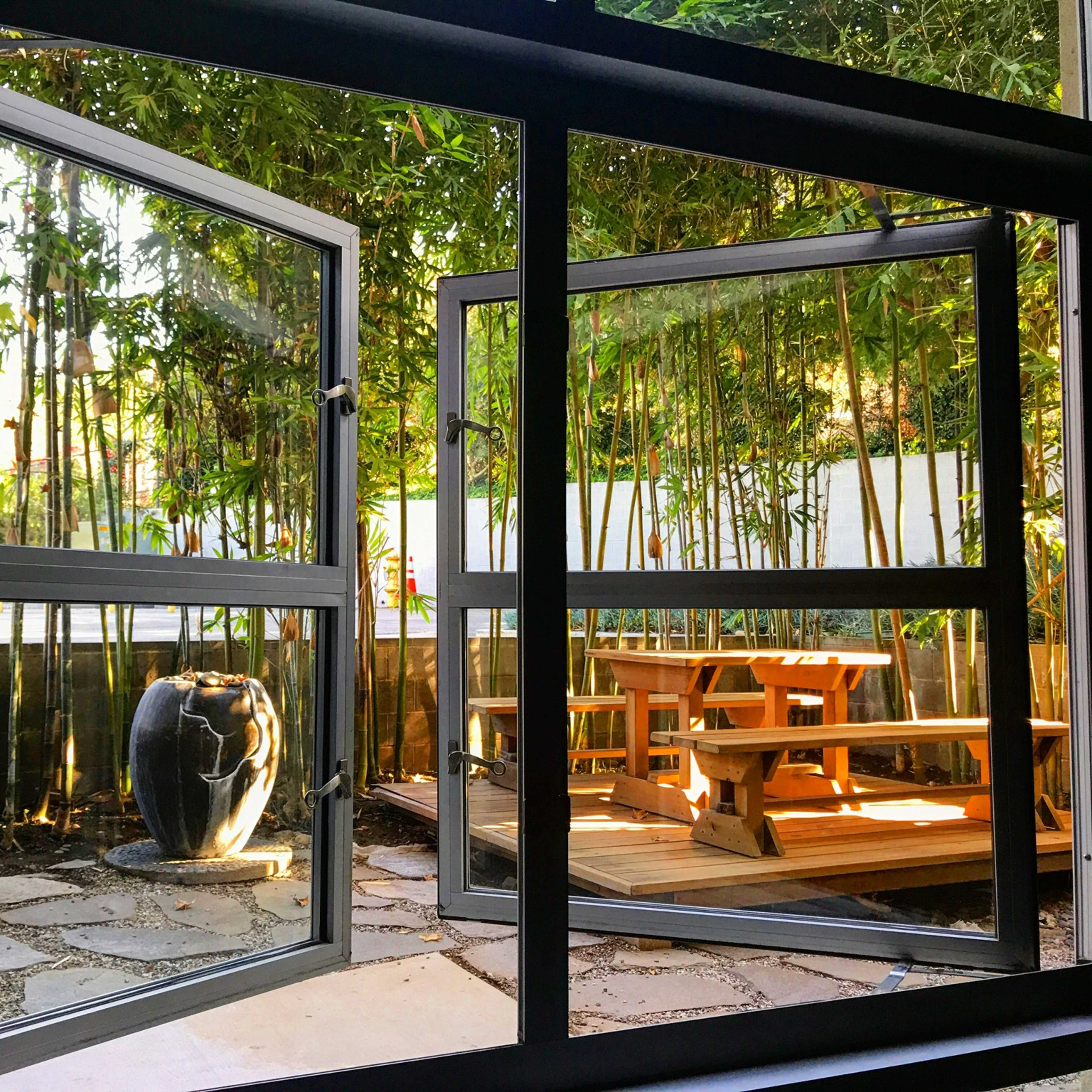 A pair of open windows frames a view of a bamboo-filled garden with a seating area and a decorative stone fountain.