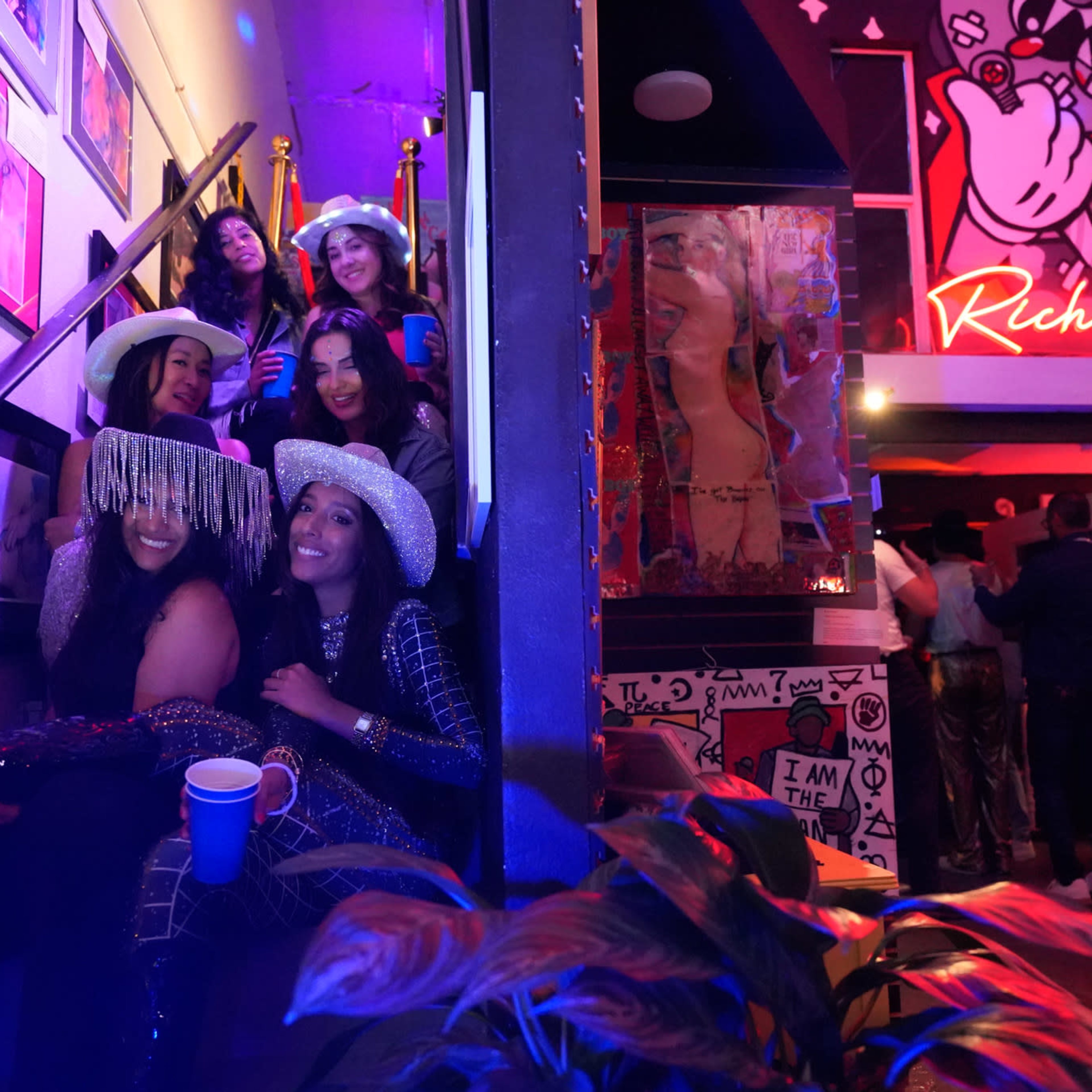 A group of women wearing hats and holding cups is seated on a staircase in a colorful venue decorated with artwork and vibrant lighting.
