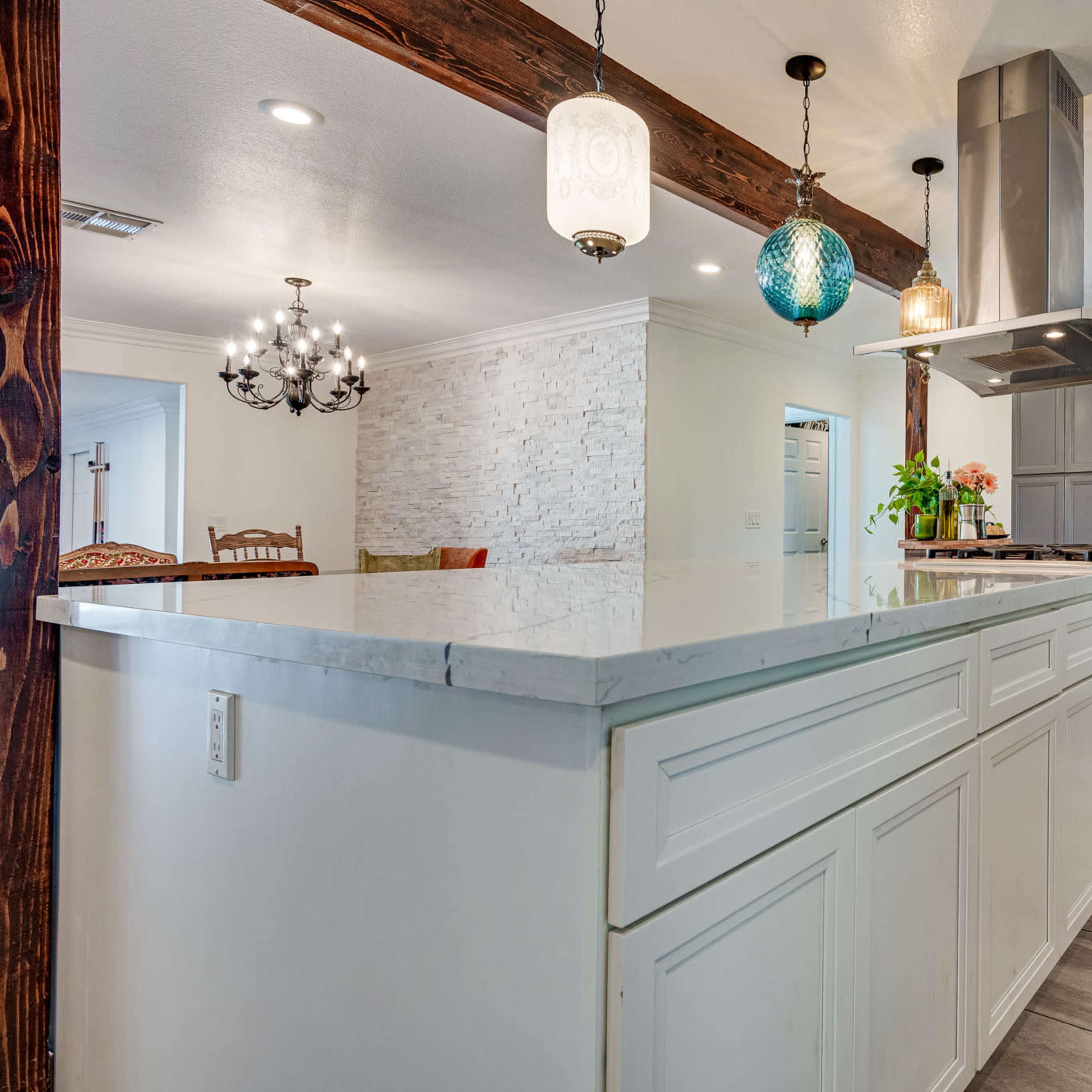 A modern kitchen with a long island counter, grey cabinetry, pendant lighting, and a stainless steel range hood.