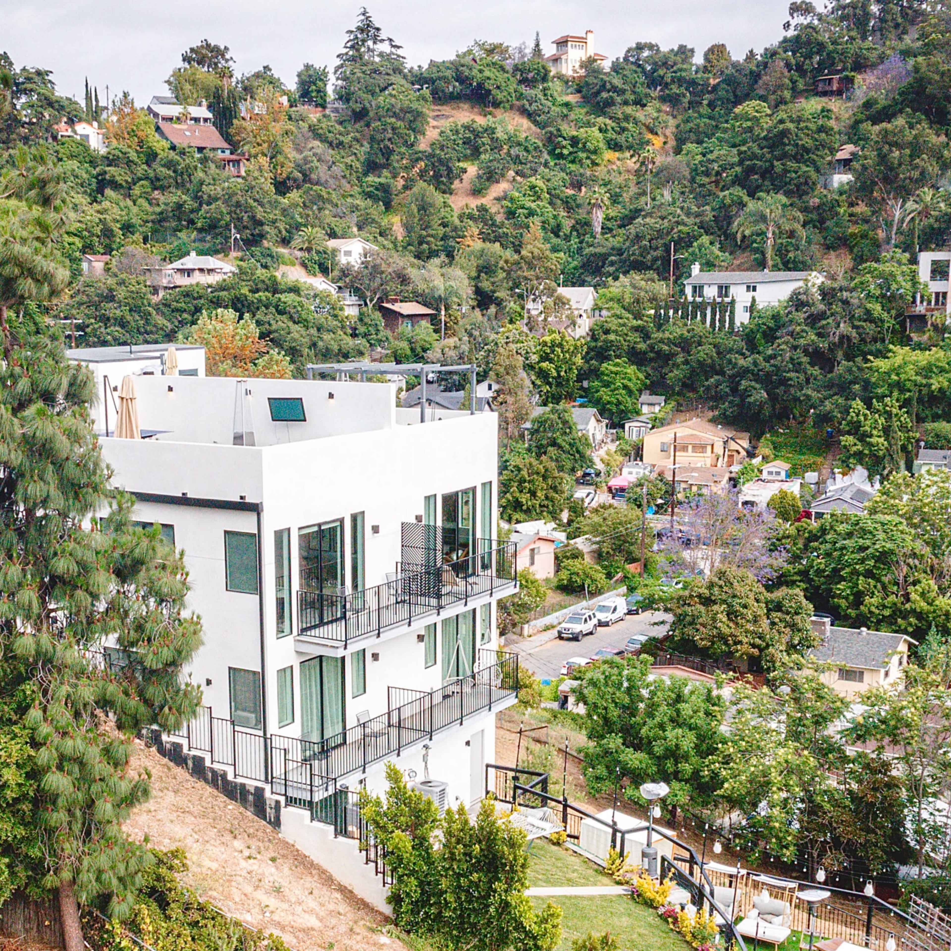 A modern white house is situated on a hillside, surrounded by lush greenery and other residences.