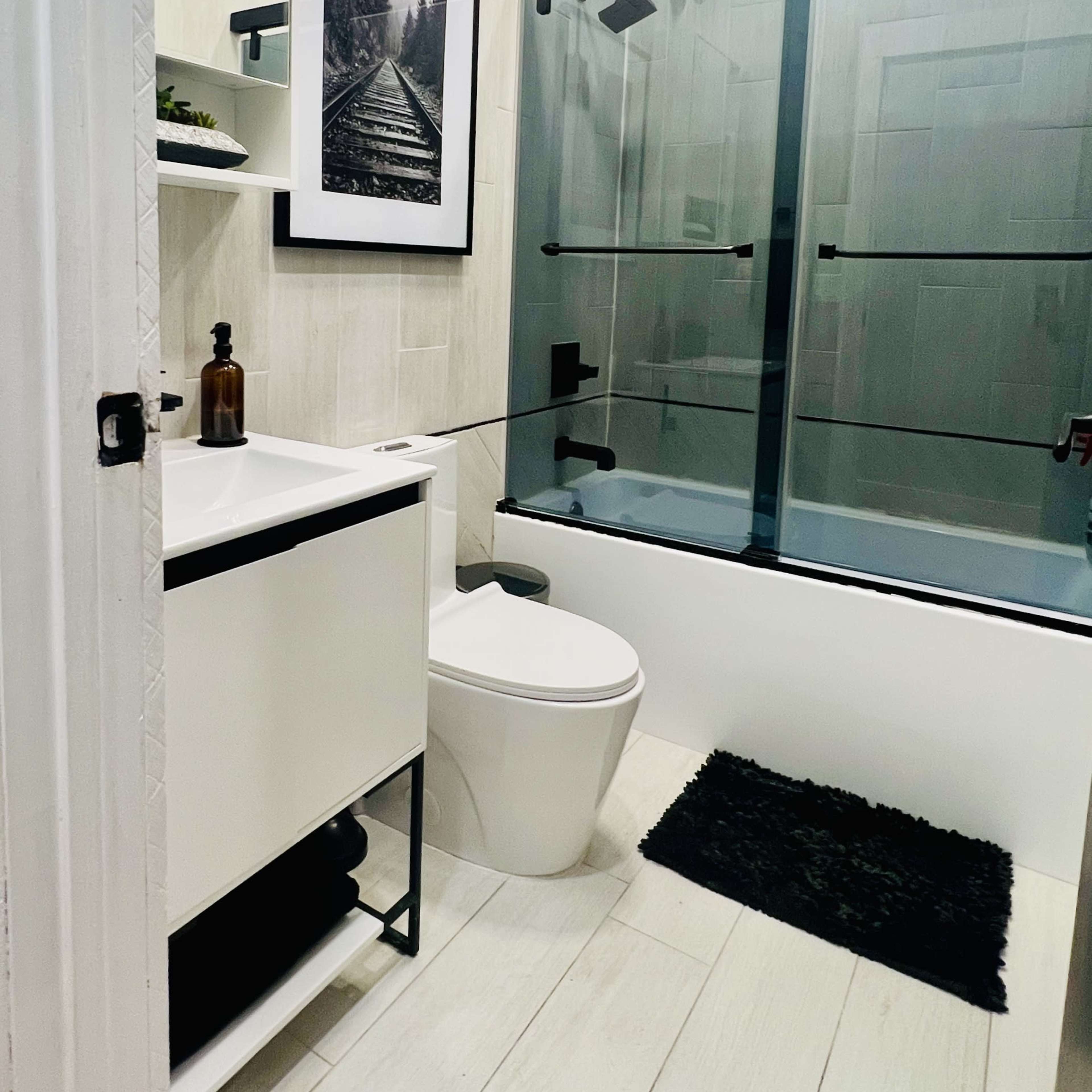 A modern bathroom featuring a glass shower enclosure, a white toilet, a sleek vanity with a mirror, and a black rug on a light-colored tiled floor.