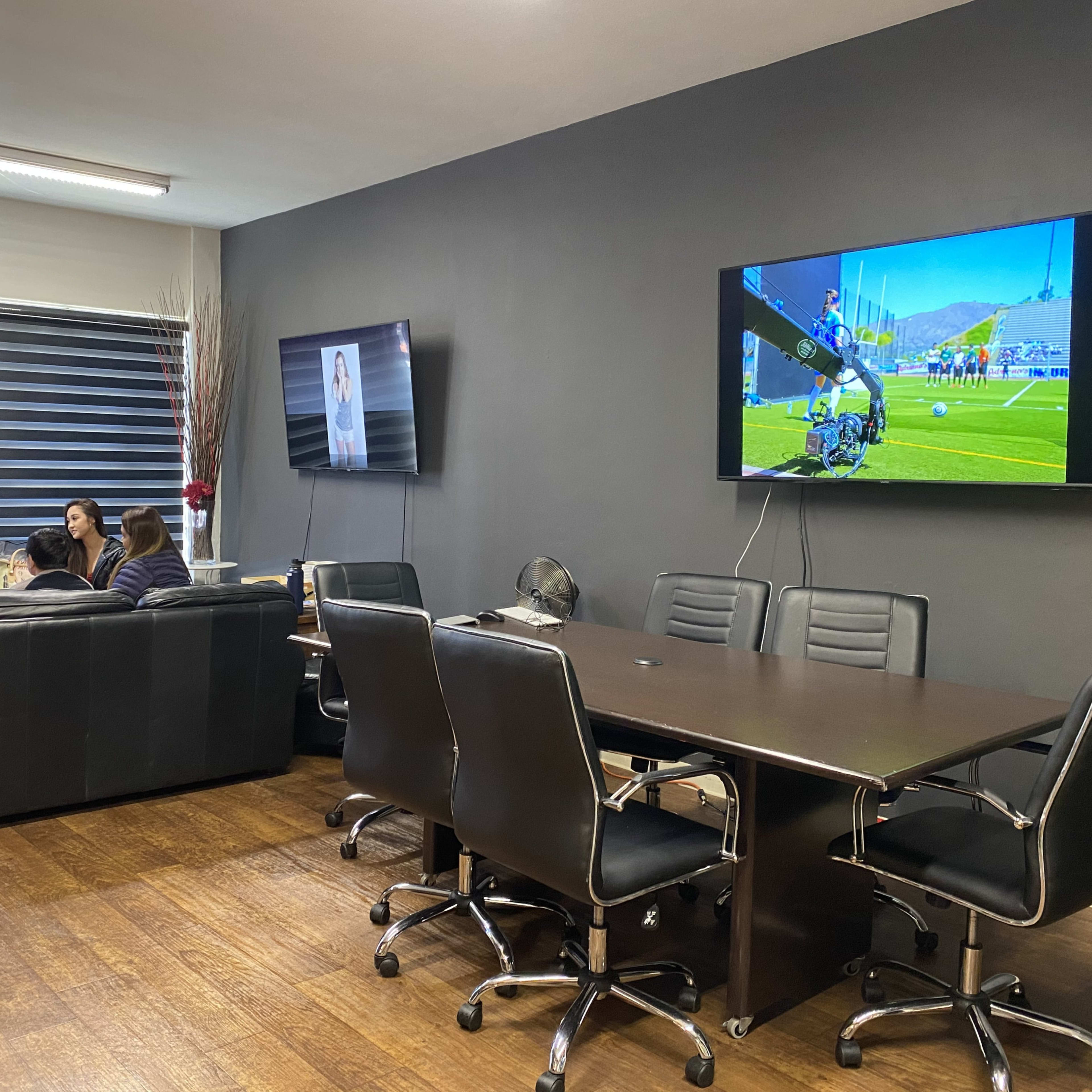 A modern office meeting room with a large table and black chairs, where three people are seated on a couch, and two screens display images on the walls.