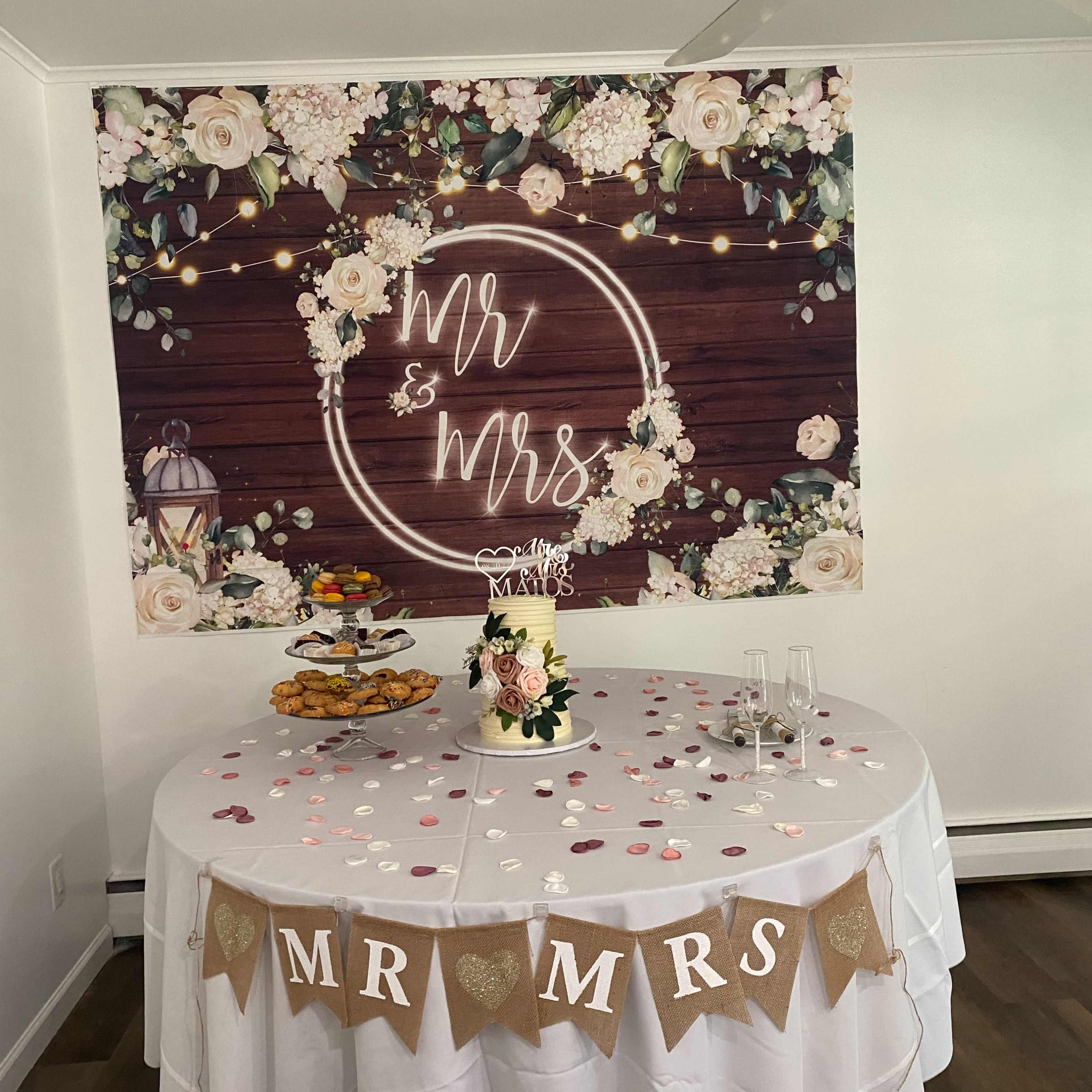 A round table decorated with a floral cake, cookies, and a banner reading "MR & MRS" is set against a backdrop featuring the same words surrounded by lights and flowers.