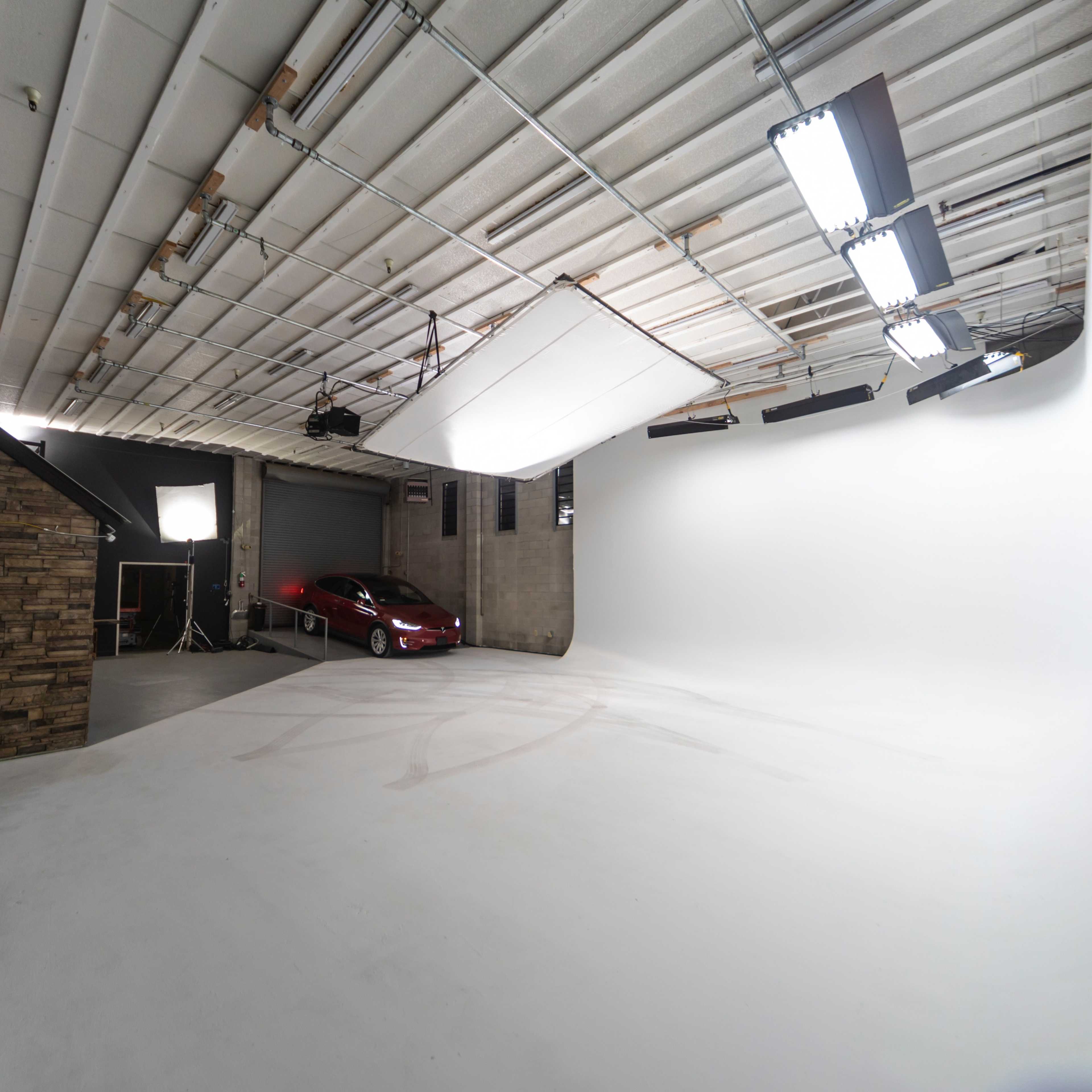 A spacious studio with a white backdrop, overhead lighting, and a car positioned in the corner.
