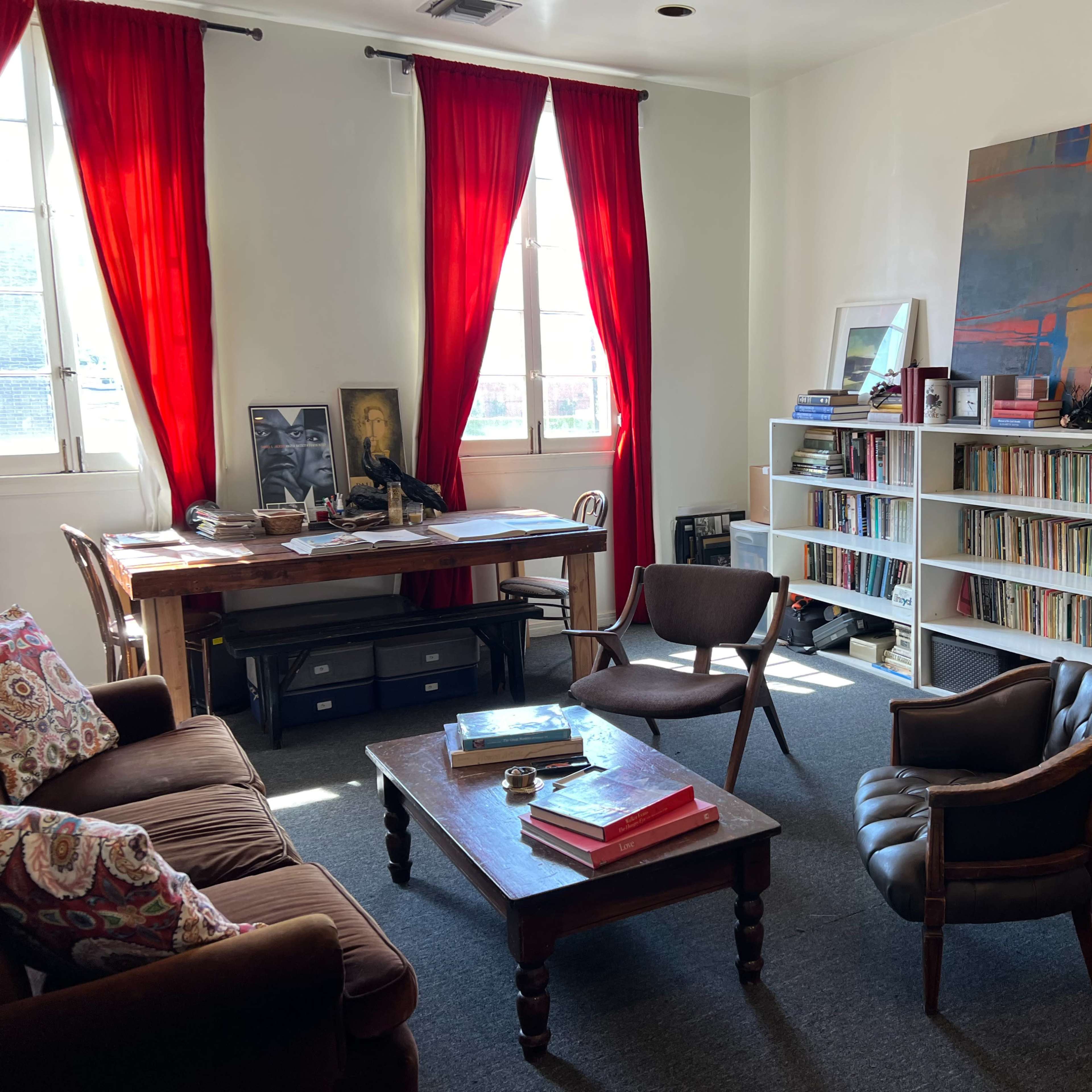 A cozy room featuring a wooden table and chairs, a bookshelf filled with records, and red curtains framing the windows.