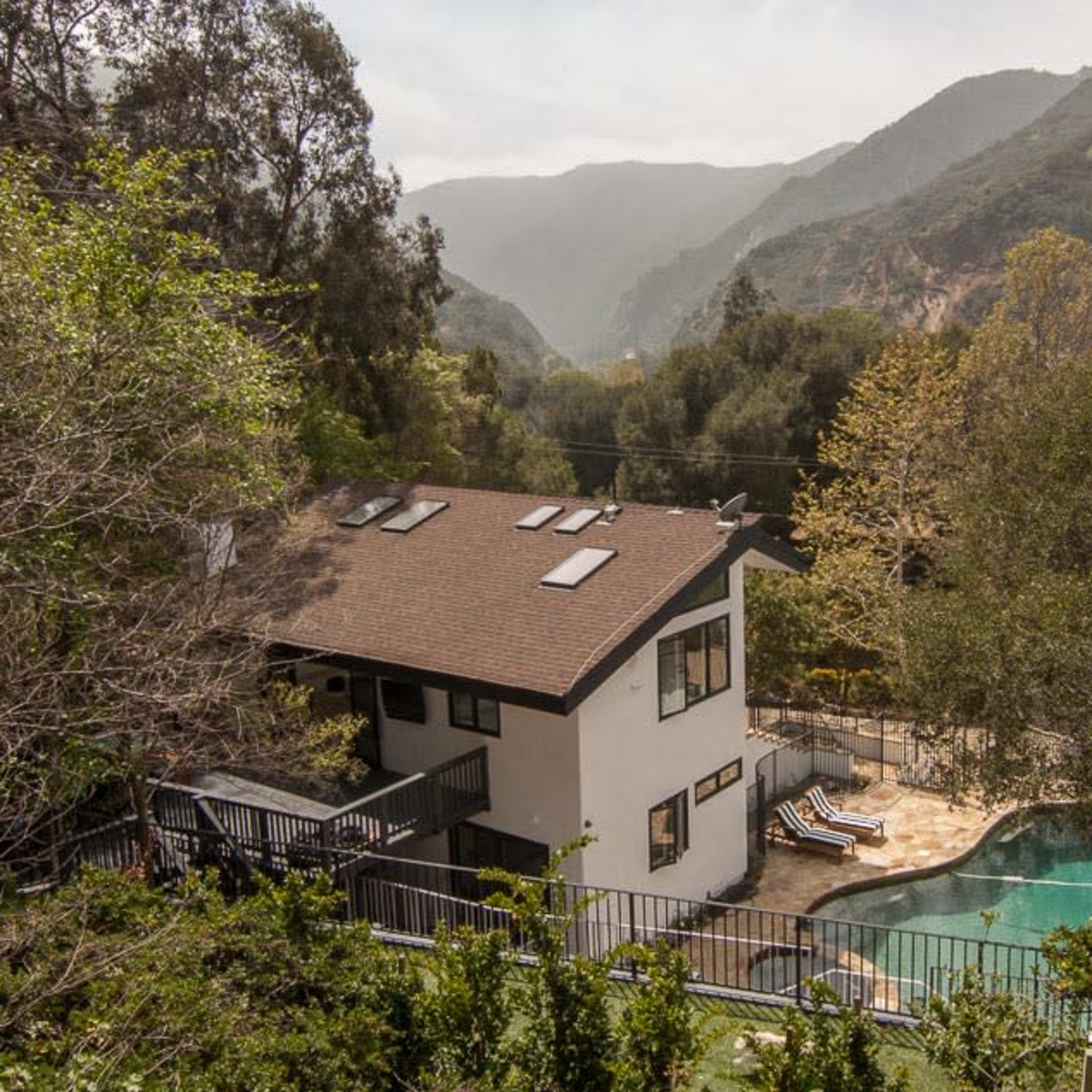 A two-story house with a pool is situated amidst a mountainous landscape surrounded by trees.