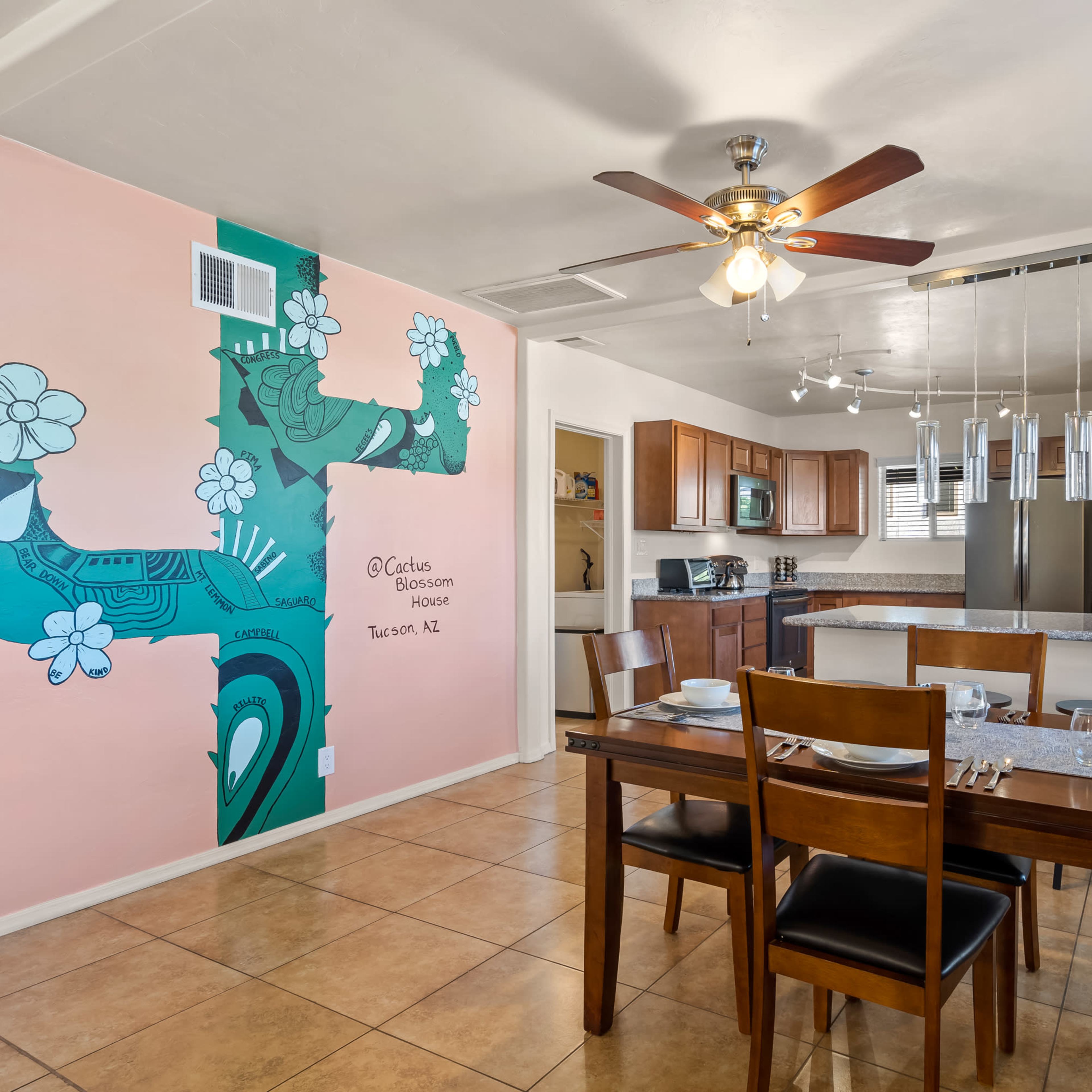 A dining area with a wooden table set for six is adjacent to a kitchen featuring warm wood cabinetry and a painted mural on the wall.