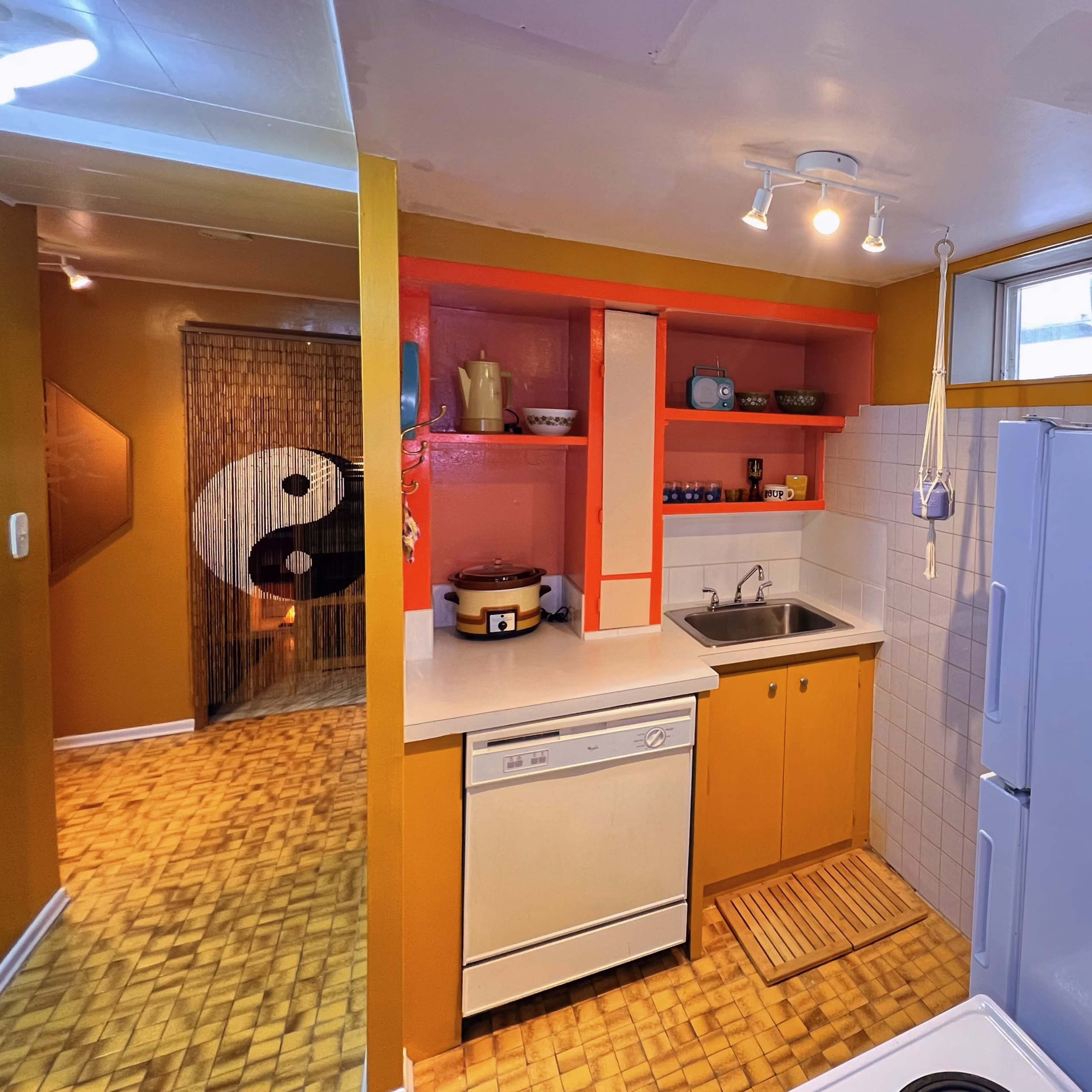 A small kitchen with orange and yellow walls, featuring a sink, dishwasher, and open shelving, leading into a room with a decorative yin-yang design.