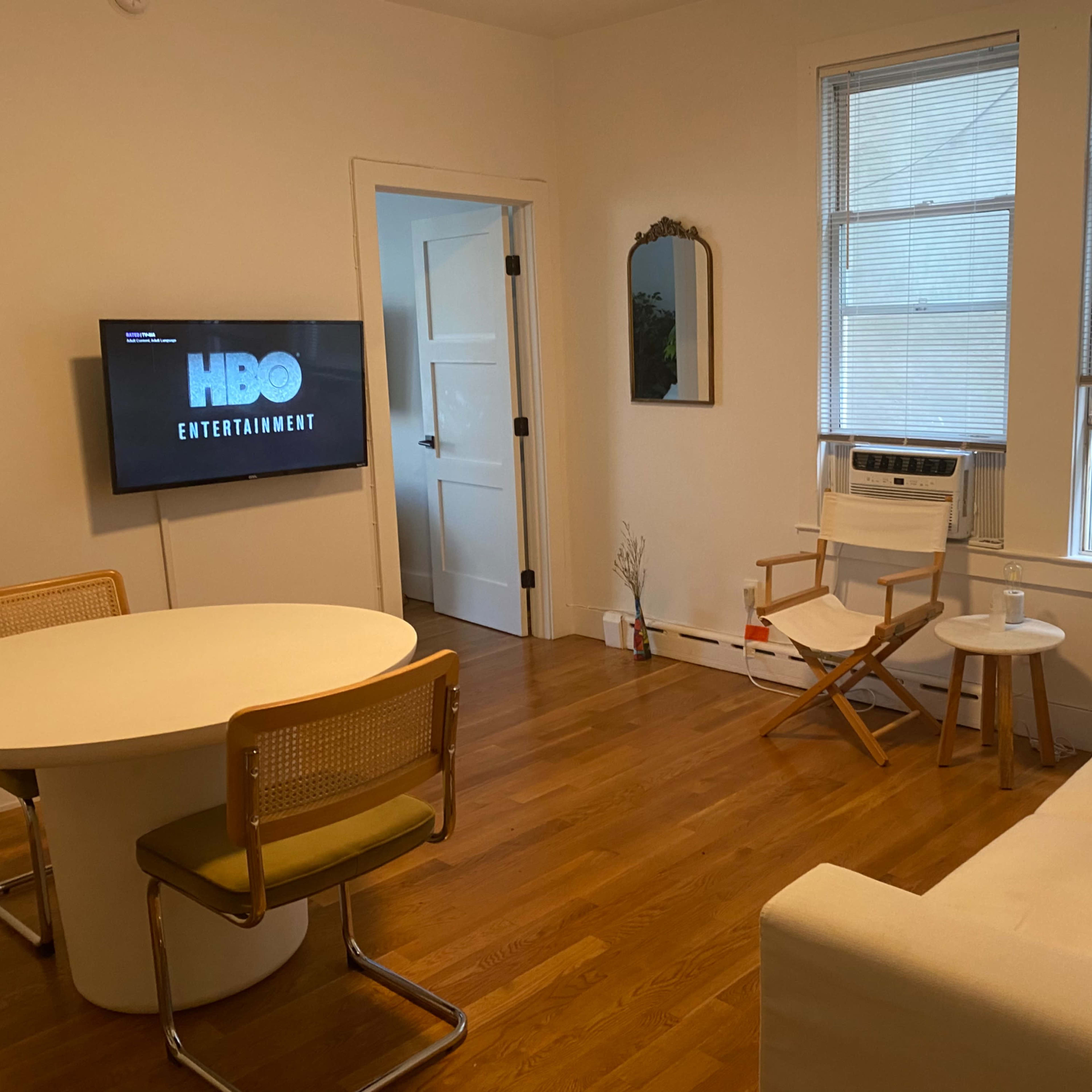 A living room with a round table, a sofa, a chair, a wall-mounted TV displaying the HBO logo, and windows with blinds.