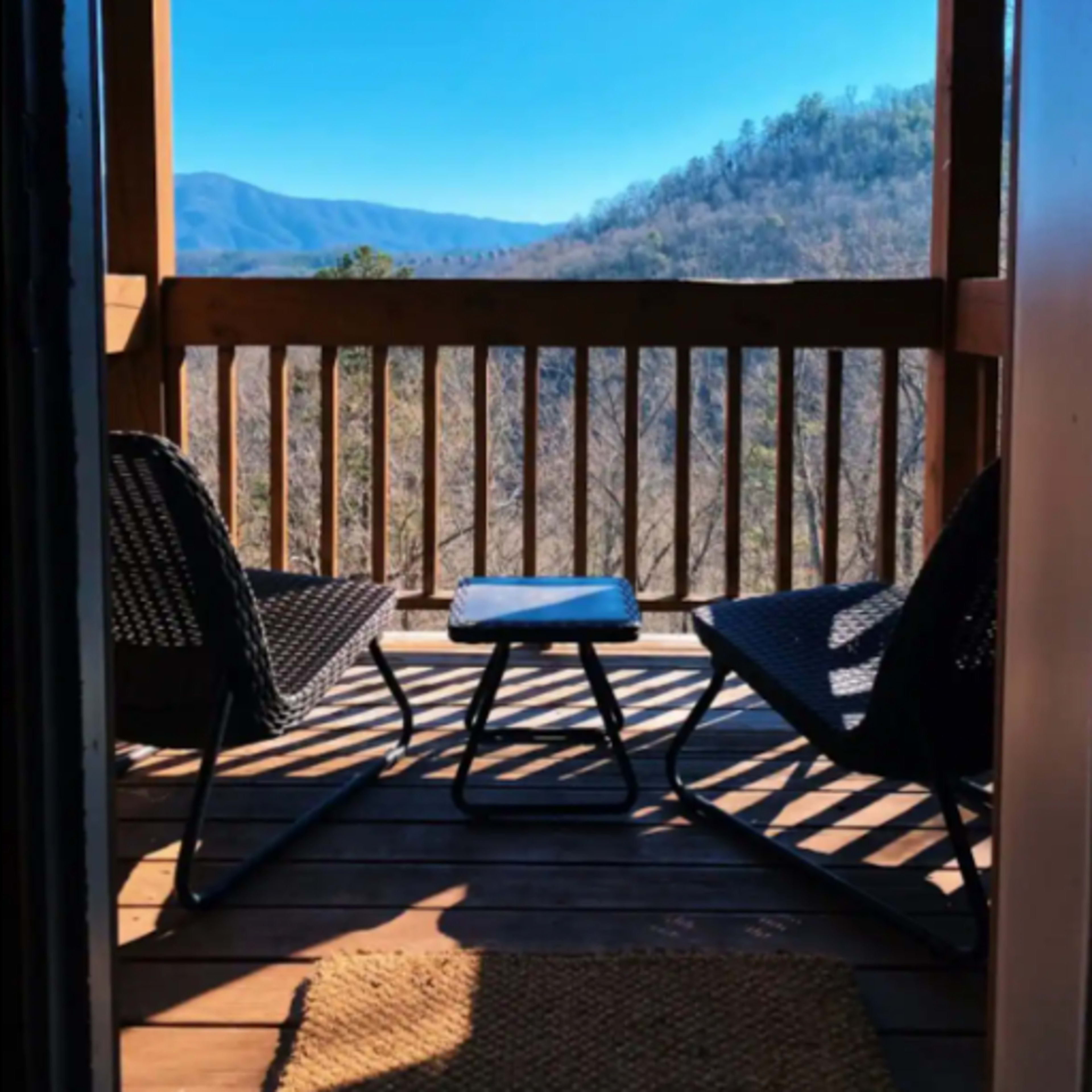 A balcony with two chairs and a small table overlooks a mountainous landscape under a clear blue sky.