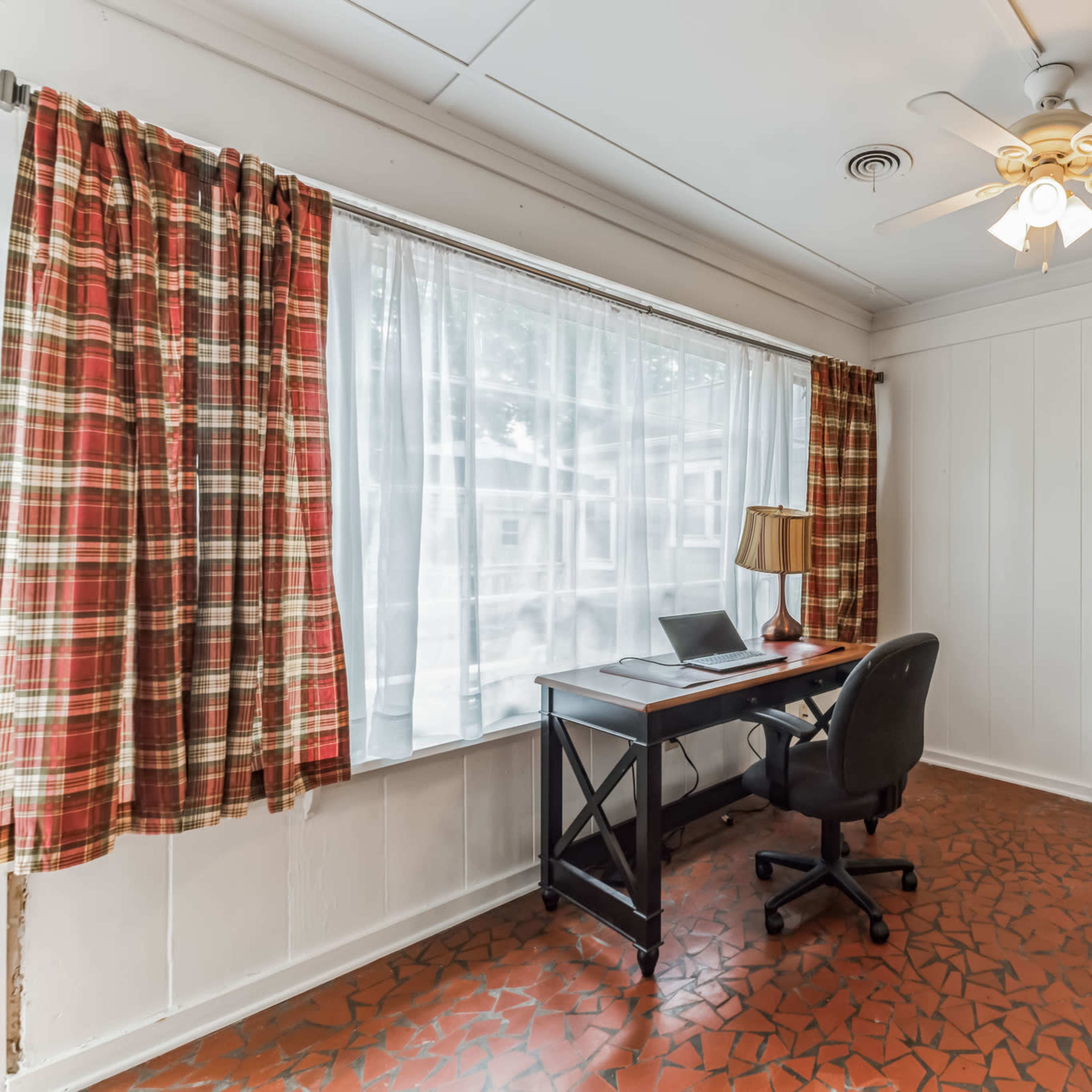 A small room with a desk and a chair, a window with checkered curtains, and a door leading outside.