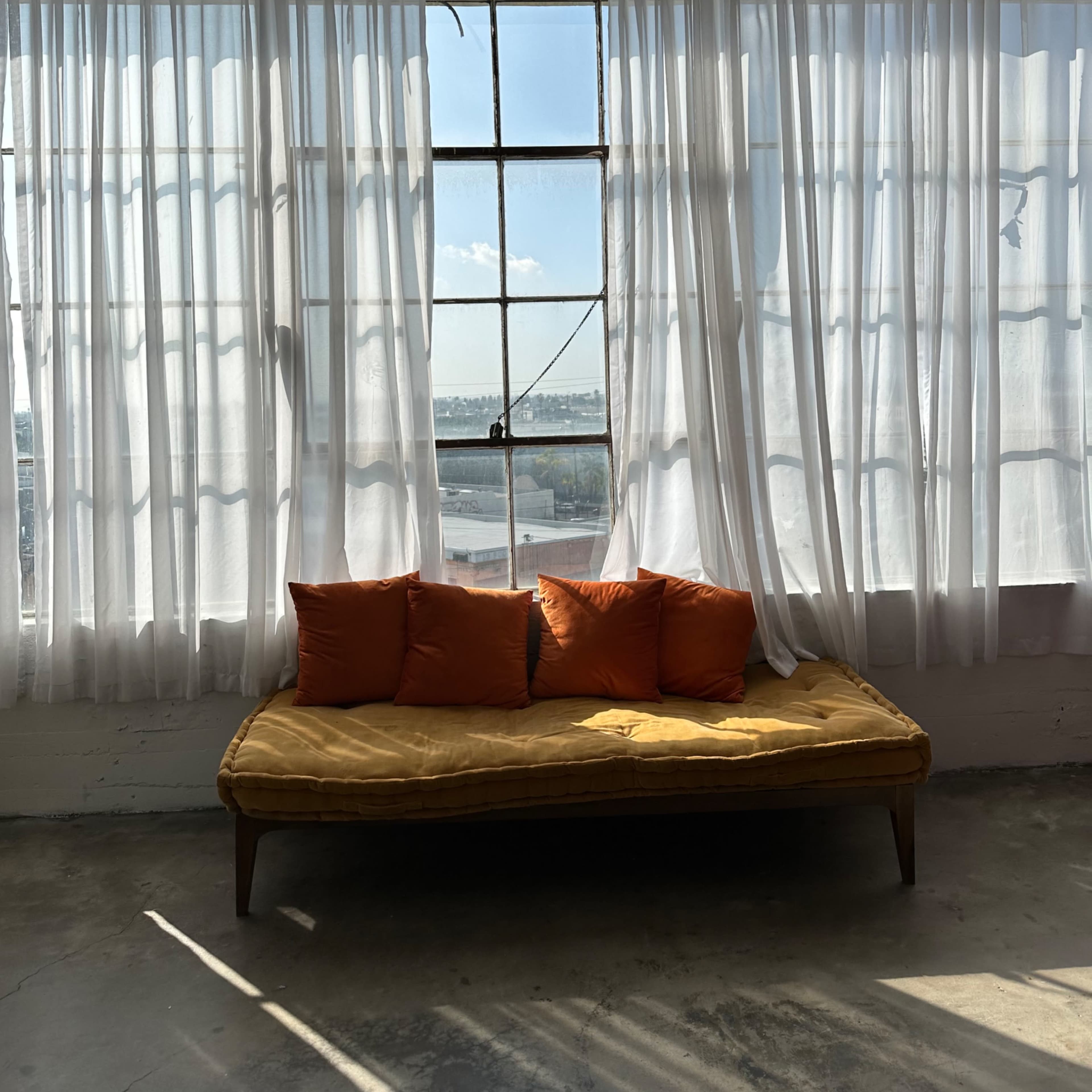 A yellow couch with four orange pillows sits against a large window adorned with sheer white curtains, allowing natural light to fill the space.