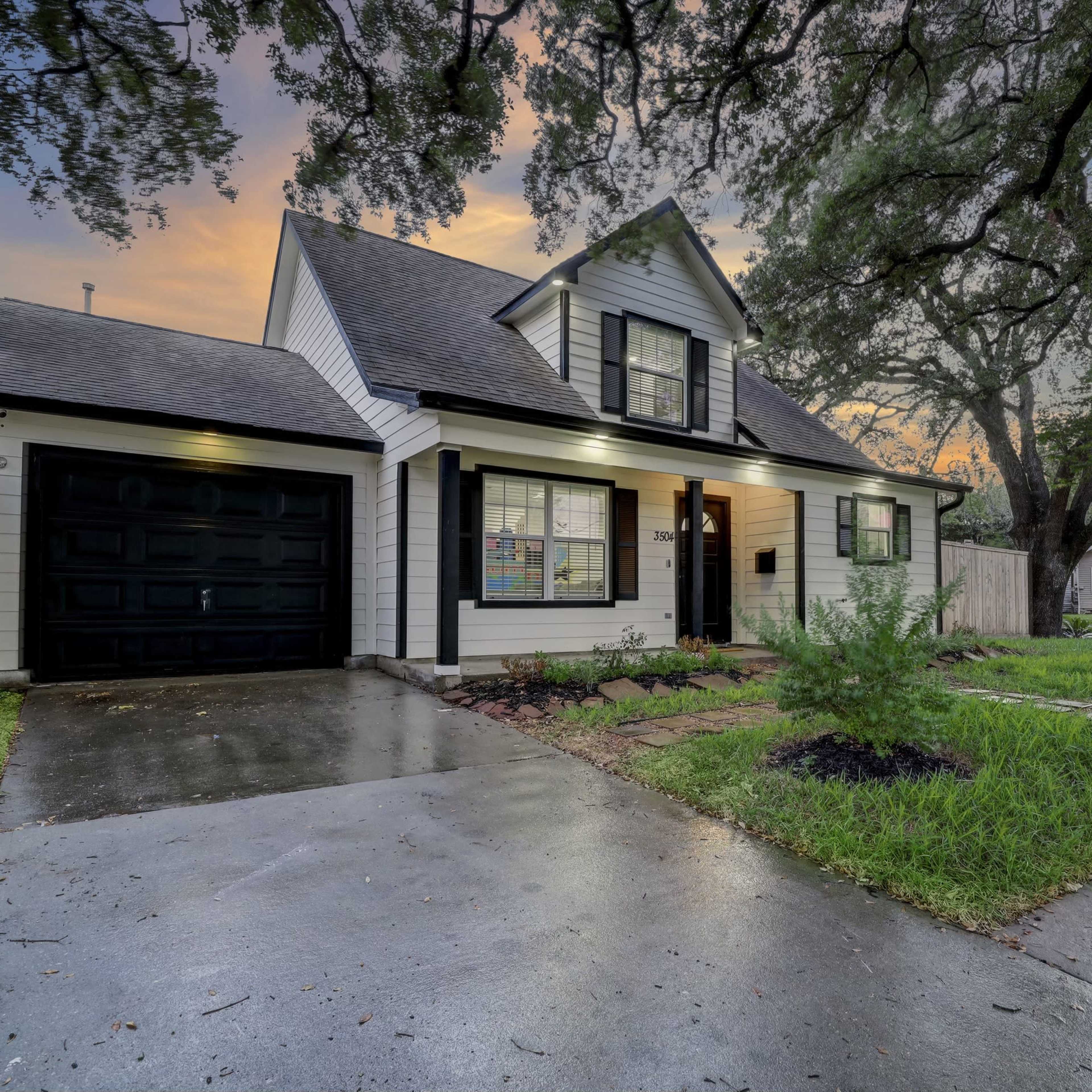 A two-story house with a gable roof, black garage door, and a stone pathway is situated on a wet driveway in a suburban neighborhood during sunset.