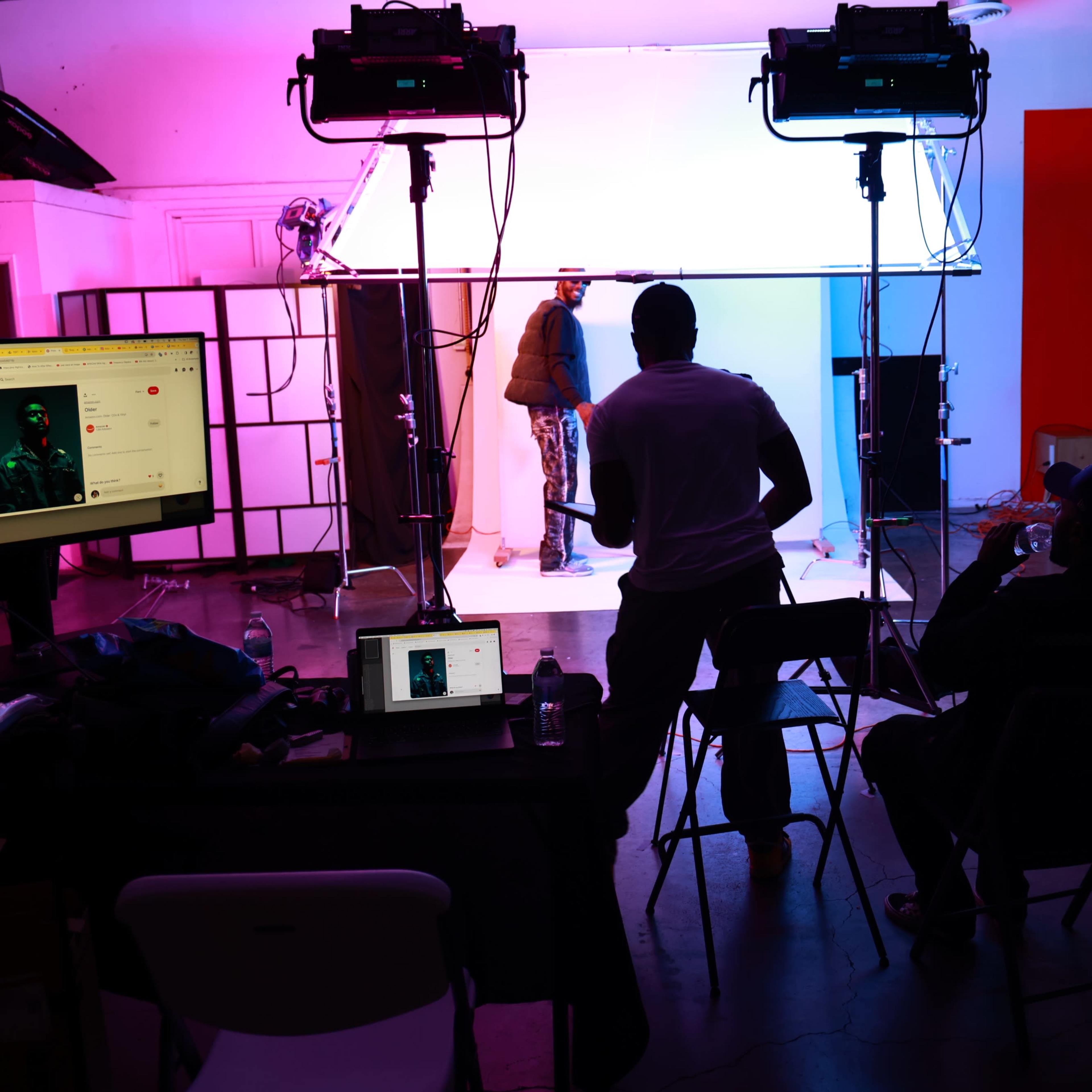 A photography studio setup features multiple lights illuminating a subject against a backdrop, with several people observing from chairs and laptops nearby.