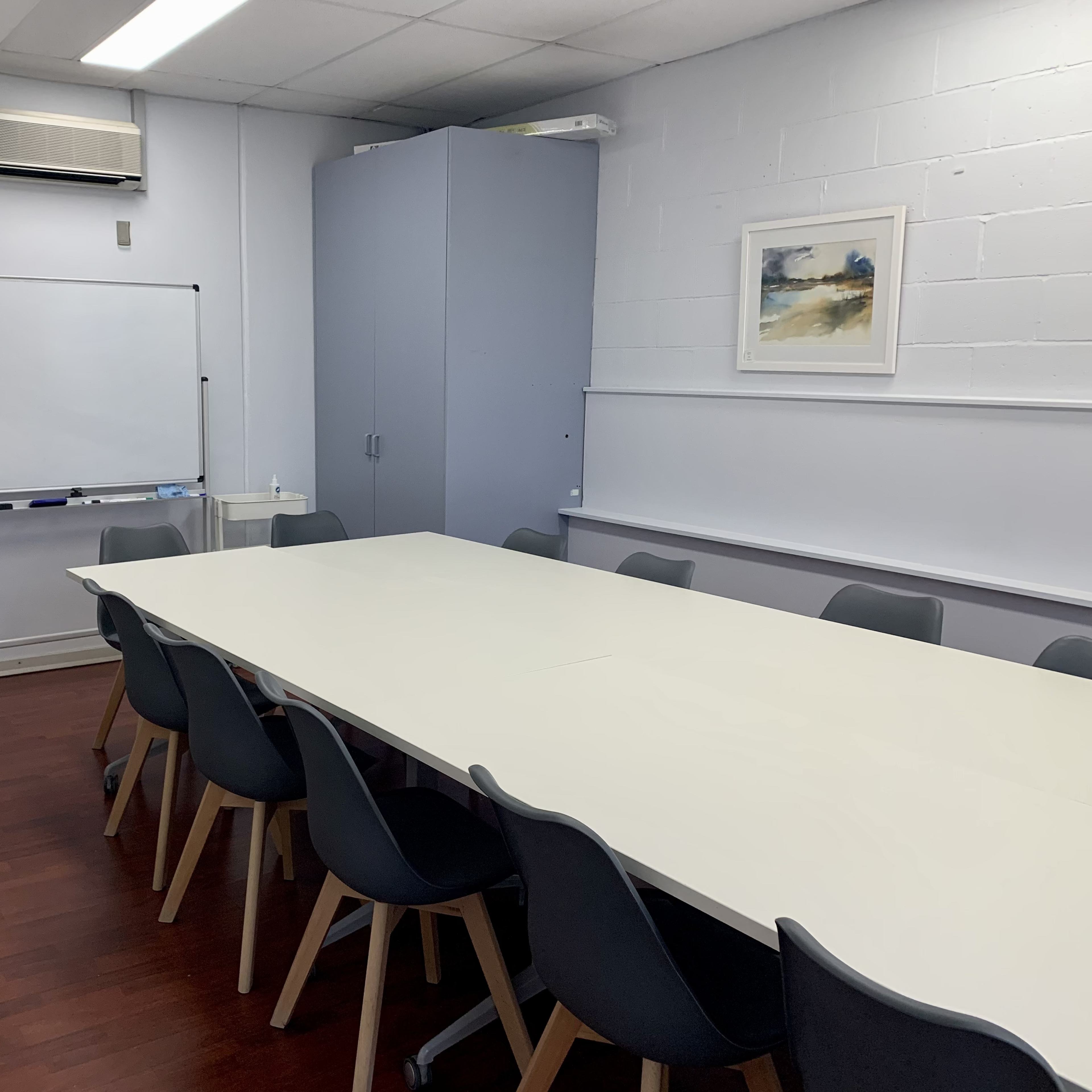 A meeting room with a large white table surrounded by gray chairs, a whiteboard, and a cabinet.