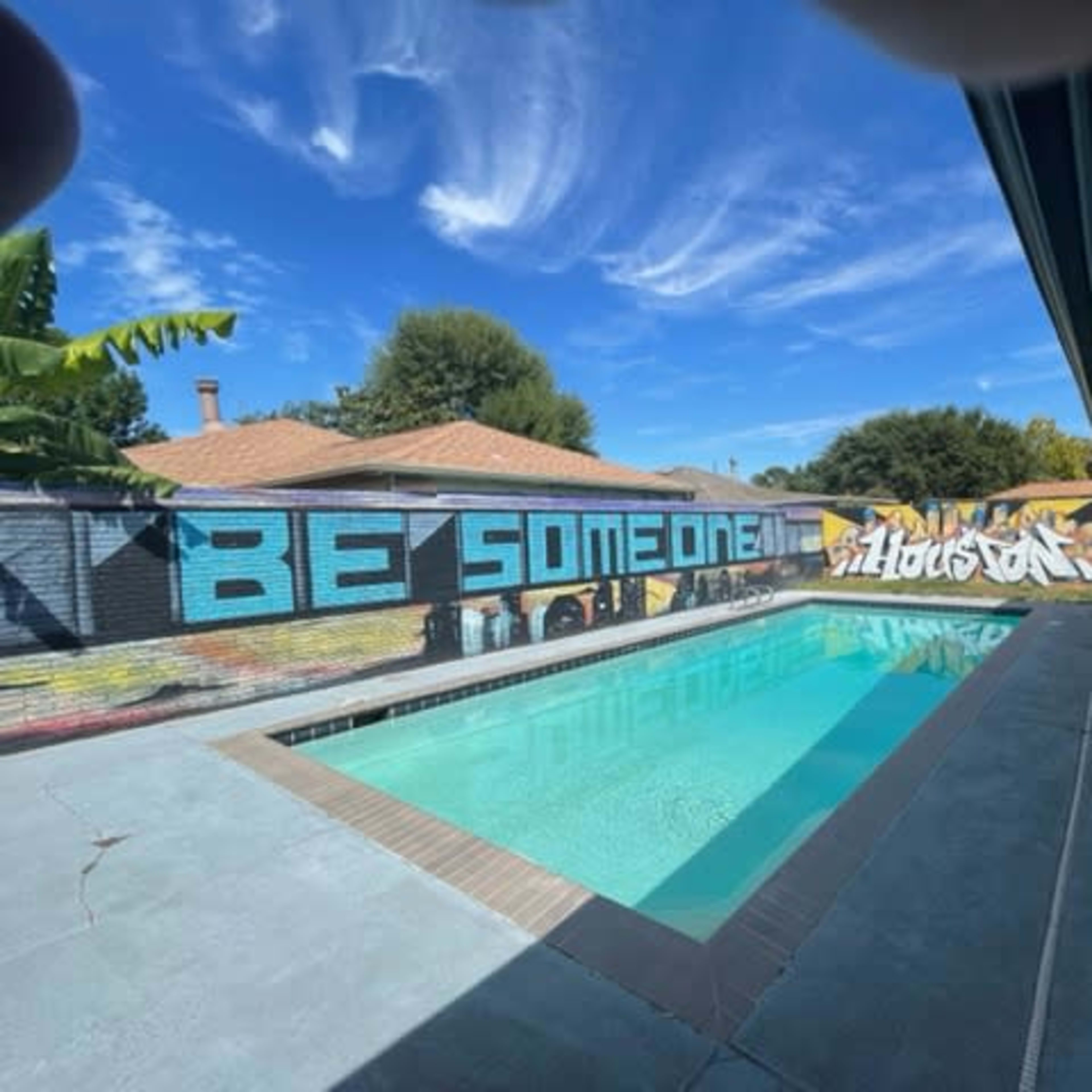 A swimming pool surrounded by a modern home, featuring a vibrant mural on the wall that reads "BE SOMEONE" against a blue sky.