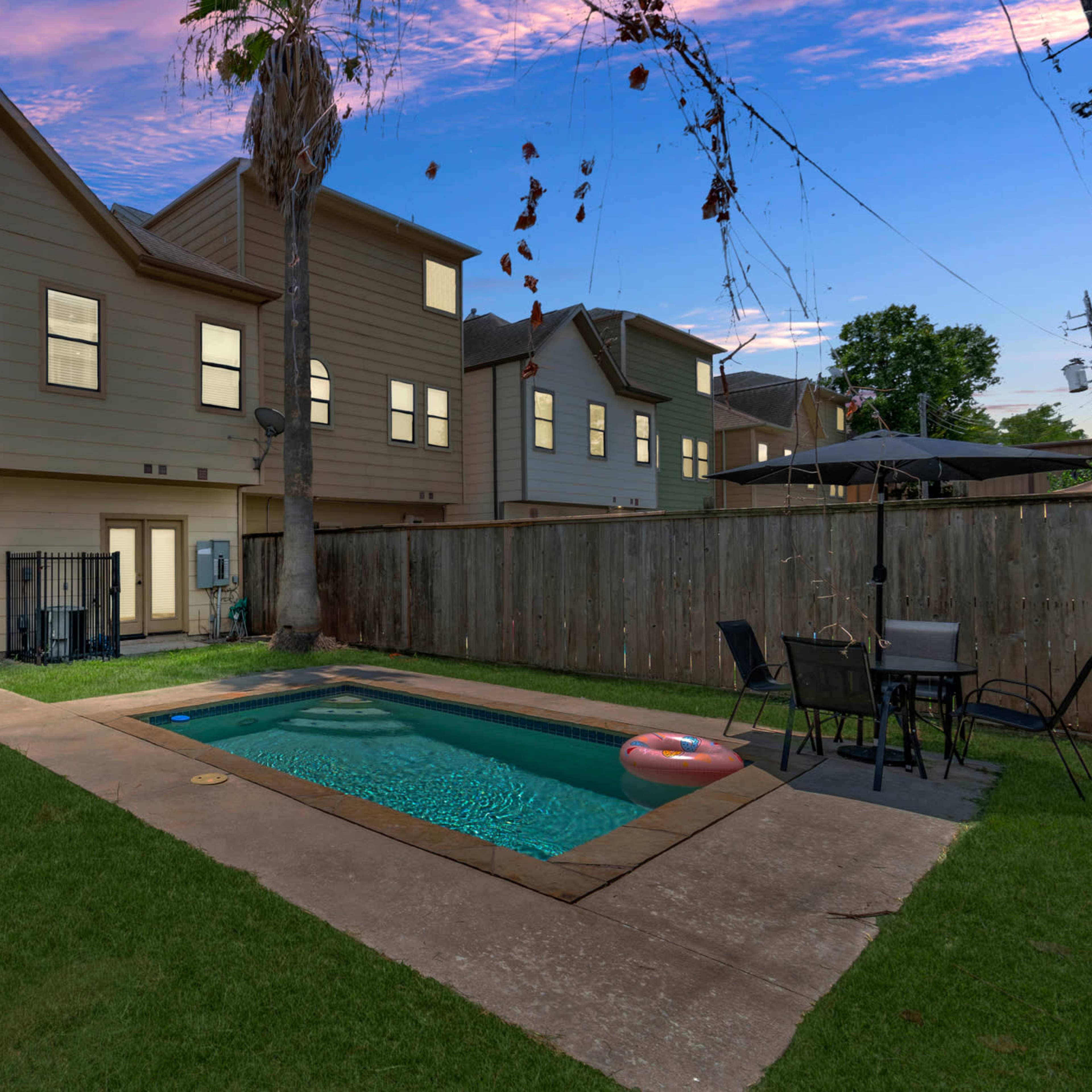 A backyard featuring a swimming pool surrounded by a grassy area, with a two-story house and a wooden fence in the background.