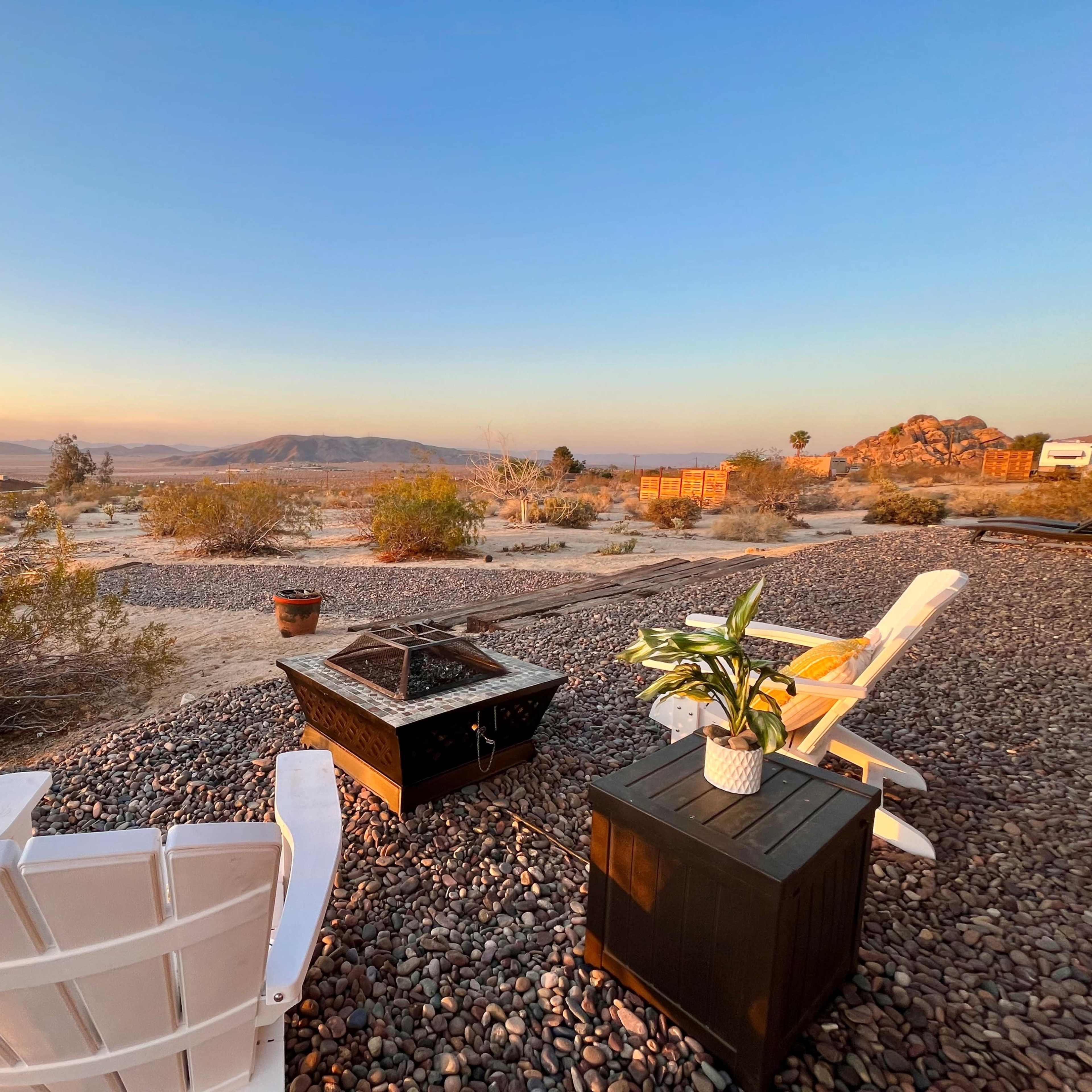 The scene shows a desert landscape at sunset with two white chairs, a black fire pit, and a potted plant on a gravel surface.